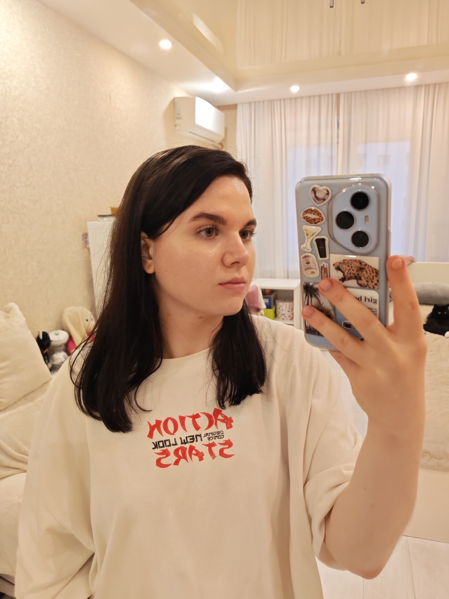 A young Caucasian woman with dark hair is taking a selfie in a room. She is wearing a cream-colored t-shirt with red text that reads "ACTION NEW LOOK STARS". She is holding up a light blue phone case decorated with stickers of lips, a cocktail glass, a coffee cup, a leopard, and palm trees. The woman is looking at her phone's reflection in a mirror. The background shows a light-colored textured wall, a white air conditioning unit on the wall, white curtains in the background, and a bed with pillows. A black cat is visible on the bed. There are no discernible features that would place the location in Bataysk, Russia.
