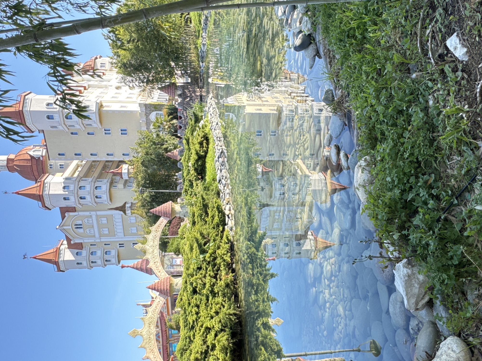 The image depicts a building with multiple towers and turrets, reminiscent of a fairytale castle, reflected in a clear body of water. The building is primarily white and tan, with red-orange roofs on the towers. The reflection is clear enough to show the architectural details of the building.
Between the building and its reflection are green bushes and trees, suggesting the presence of landscaping. The foreground of the reflection shows rocks and pebbles at the water's edge. There is also green grass and foliage near the rocks.
The image seems to be taken during a bright sunny day, with a clear blue sky visible above the building. There are no people visible in the image.
Based on the architecture, the building is likely part of the Sochi Park, which is a theme park located in Sochi, Russia. The detailed nature of the structures suggests it is a purpose-built theme park attraction.
