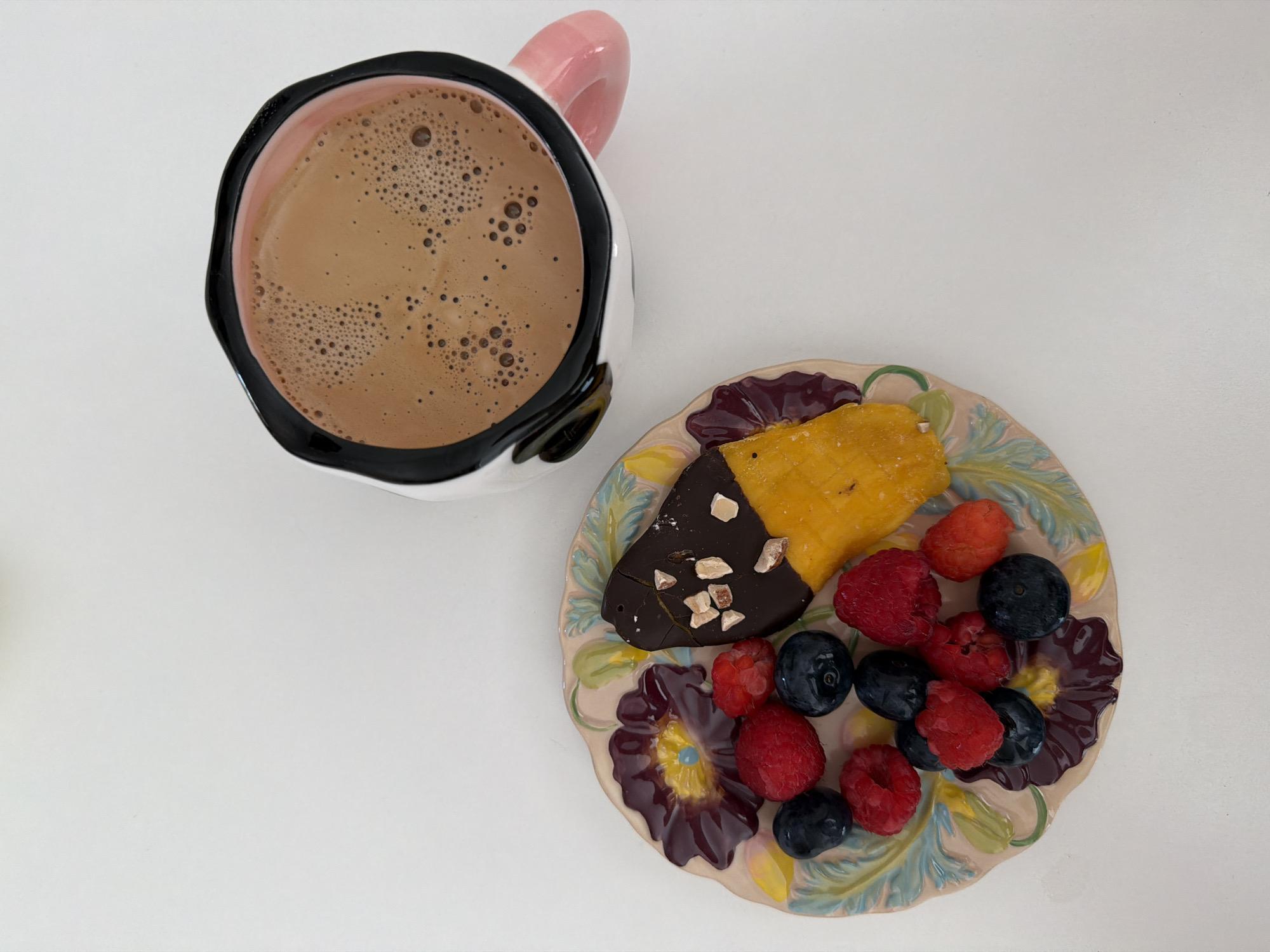 A high-angle shot shows a mug of coffee next to a plate of food. The mug is white with a black rim and a pink handle, containing a light brown liquid with foam bubbles. The plate features a piece of mango partially dipped in dark chocolate with nut pieces, and a cluster of raspberries and blueberries. The plate is decorated with floral patterns. The background is a plain white surface. There's no indication of Tel-Aviv, Israel, or any people.