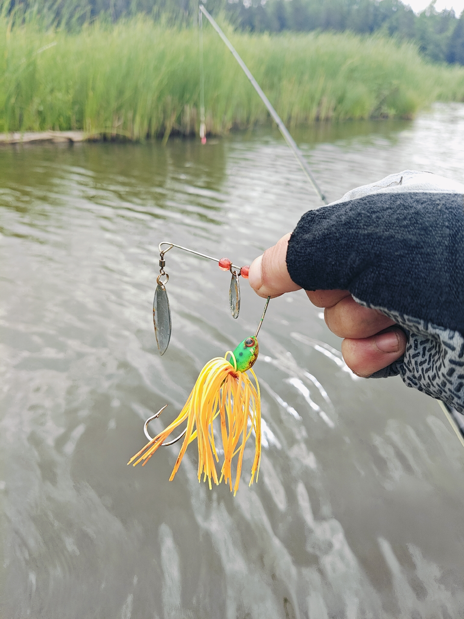 A person wearing a fishing glove holds a fishing lure with yellow skirt and a hook, over the surface of a body of water. The lure is attached to a spinner with two silver blades and red beads. The water has ripples and reflections. In the background, tall green grass lines the edge of the water, and a fishing rod is visible. The scene appears to be in a natural, outdoor setting, possibly near Detskii Dom (Bashenino), Russia, given the presence of water and vegetation. The image is a close-up shot, focusing on the lure and the gloved hand.