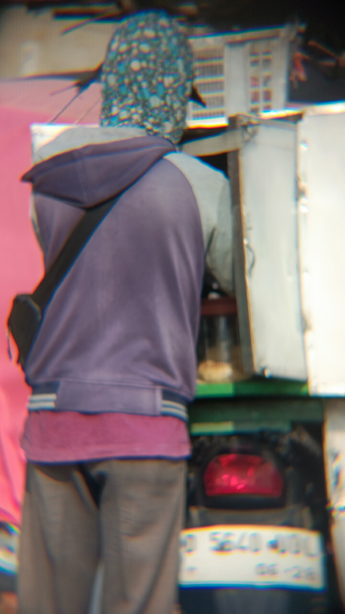 The image shows the rear view of a person wearing a purple hooded jacket over a pink shirt and gray pants. The person has a black strap across their chest, suggesting they are carrying a bag. They are standing in front of a vehicle that has a red taillight and a license plate that appears to be partially visible. The background includes a structure with windows and a pink surface on the left. The overall image quality is distorted with chromatic aberration, particularly around the edges and in the details. The scene appears to be an outdoor urban environment, possibly near a street or roadside. The vehicle's license plate suggests it is registered in Indonesia.