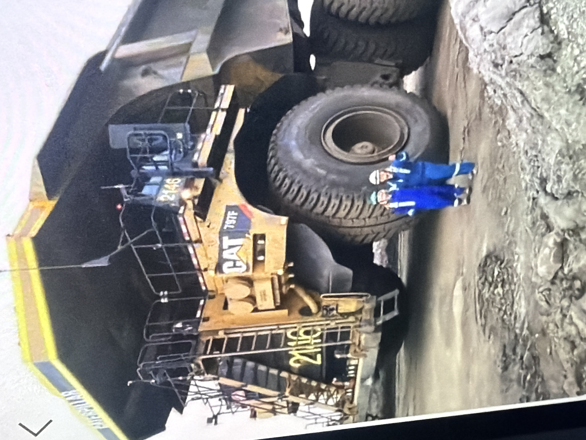 The image captures a close-up view of a massive yellow Caterpillar mining truck, identified as a CAT 797F. The truck's large wheels and immense size are emphasized, dwarfing two individuals standing in front of it. These individuals are wearing blue overalls and hard hats, suggesting they are workers on the mining site. The truck is parked on a dirt or gravel surface, indicative of a mining or industrial environment. The background is blurred, but the overall scene suggests an operational mining location. The text "CATERPILLAR" is visible on the upper part of the truck, along with the model number "797F" and "2146". The number "2116" is prominently displayed on the side of the truck's body. The location is specified as the city of Bukuru, Nigeria.