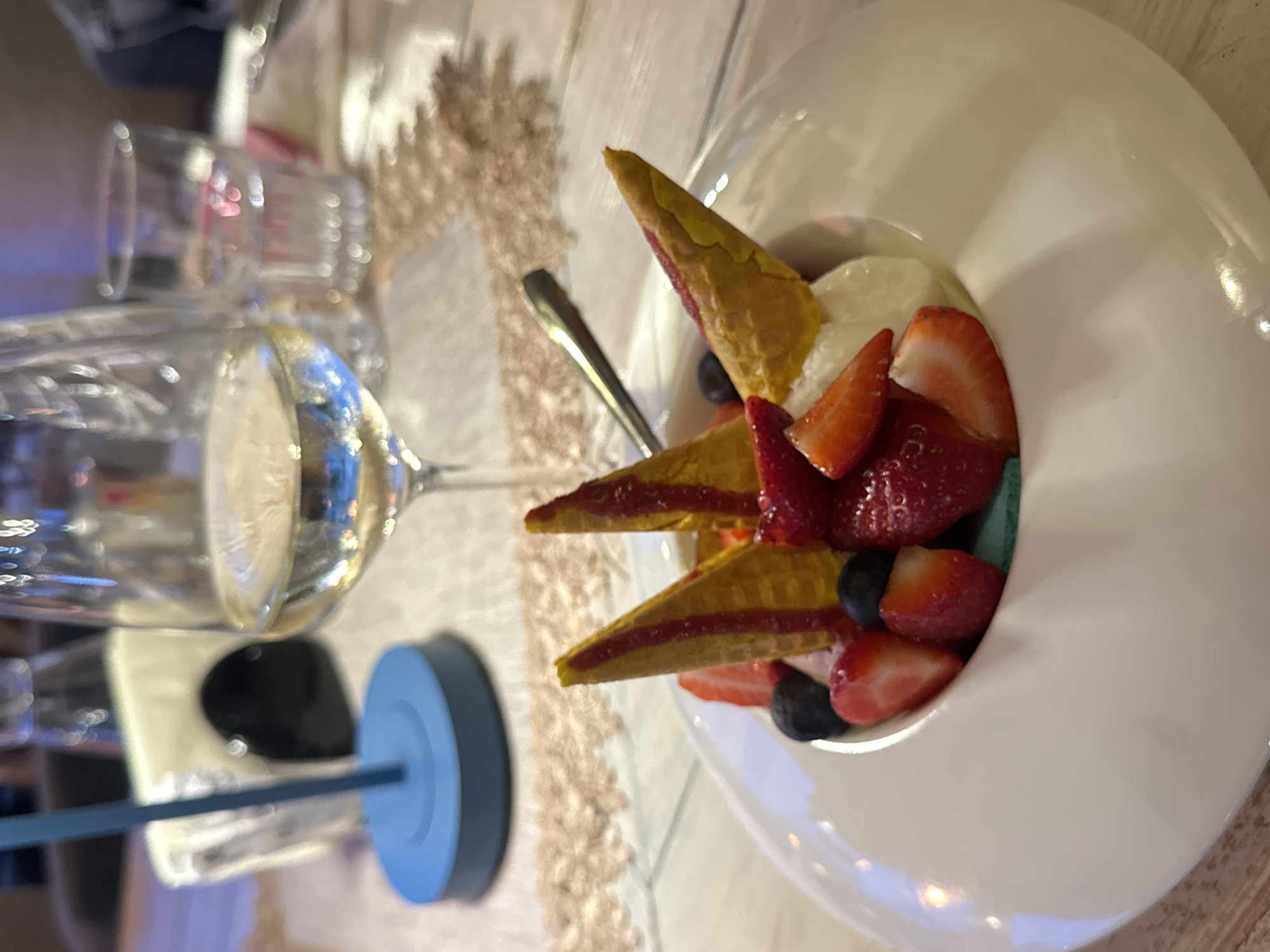 The image depicts a table setting in Veliko Tarnovo, Bulgaria. A partially filled wine glass with a blue straw and lid sits beside a white bowl of dessert. The dessert consists of ice cream, strawberries, blueberries, and cone-shaped pastries. A spoon rests in the dessert bowl. A patterned tablecloth or placemat is visible beneath the objects.