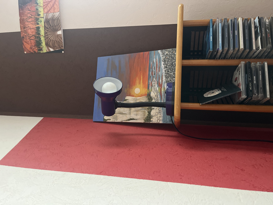 The scene is situated in a room in Ancenis, France. The image captures an indoor corner where two walls meet. The left wall features a textured white surface partially covered by a diagonal, textured red paint stripe. The right wall is divided vertically, with a darker brown textured section on the left and a lighter beige or off-white textured section on the right.
A light-colored wooden shelf unit is positioned in the corner. On the top surface of this unit, a purple desk lamp with a flexible black neck and a white interior reflector is present, illuminating a visible light bulb. To the right and behind the lamp, an unframed rectangular painting depicts a sunset over a body of water with prominent white cliffs on the left, a bright orange sun setting over the horizon, and a foreground of grey pebbles or rocks.
Higher on the brown section of the right wall, a horizontally oriented framed or mounted picture is displayed. This picture appears to be a diptych or collage, with the left half showing autumn trees with red foliage and the right half displaying an architectural or geometric pattern with circular elements in earth tones.
On the lower shelf of the wooden unit, two stacks of compact disc (CD) cases are visible. The left stack includes a partially obscured case with a white circular graphic on a dark background, and cases labeled "AC/DC Black Ice" and "The Doors Strange Days." The right stack is taller, with several CD cases displaying blue or light-colored spines, including "Joe Satriani - Crystal Planet" at the top. A black power cord for the desk lamp extends downwards along the left wall. No individuals are present within the frame.