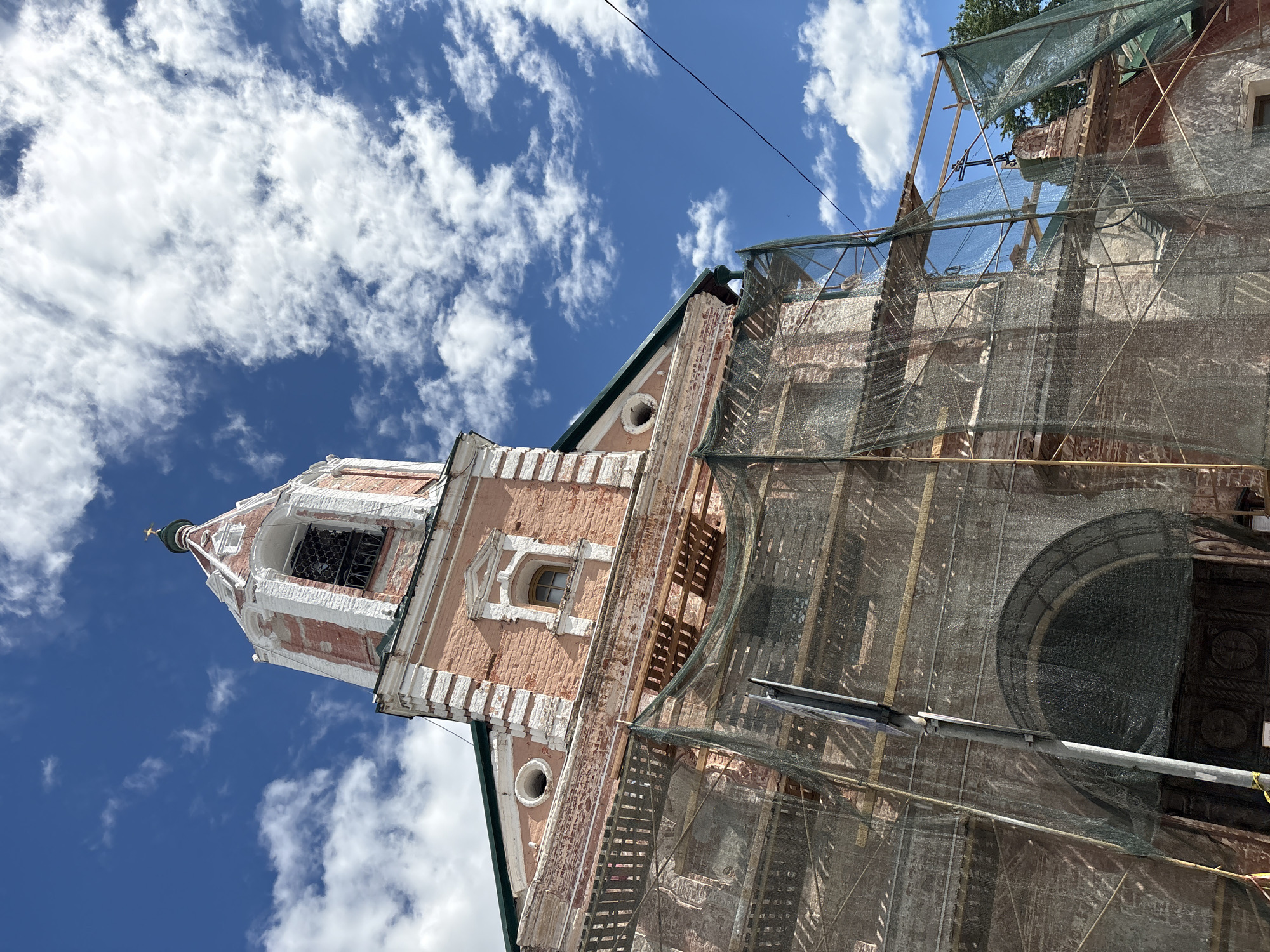This image shows the bell tower and part of the main building of a church in Pereslavl-Zalessky, Russia, under renovation. The building's facade is made of pink brick with white decorative elements. The bell tower has a white spire with a golden cross. The main building is partially covered with scaffolding and a green mesh net, suggesting ongoing construction or repair work. Wooden beams and planks are visible through the netting. A small arched window is visible on the pink brick section of the building. The sky is bright blue with scattered white clouds. The sun is shining, indicating it is daytime. The overall scene is of historical architecture undergoing preservation. No people are visible in the image. There is no visible text.