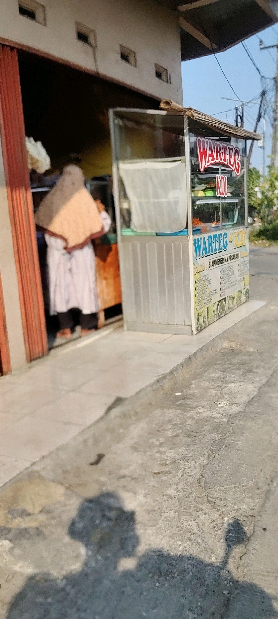 In Mekarmukti, Indonesia, a street food stall with a glass display case is positioned next to an open doorway of a building. The stall, identified by the sign "WARTEG KDI," displays various food items within the glass case. The building's facade features a textured light-colored wall with several small rectangular openings. Red corrugated metal doors are partially open to reveal the interior, where a person wearing a light-colored dress and a patterned head covering is partially visible. The ground in front of the stall and building consists of paved tiles transitioning to a concrete sidewalk and road. Sunlight casts distinct shadows of people and a motorcycle onto the ground. The background shows a clear blue sky with visible utility poles and wires, indicating an outdoor urban or semi-urban setting.