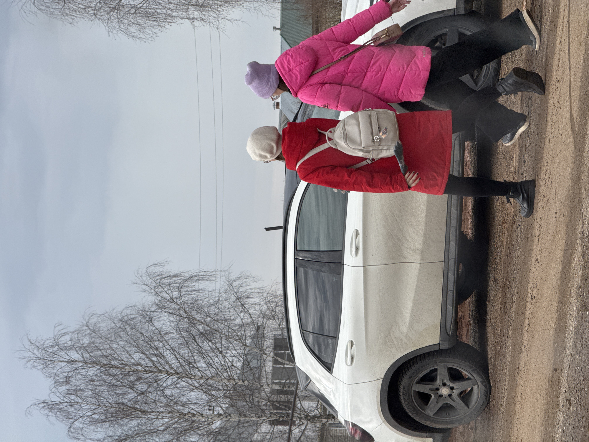 Two individuals are depicted outdoors near a white vehicle, possibly a car or SUV. The person closest to the camera is wearing a red coat, black leggings, and black boots, with a light-colored beret and a light-colored backpack. The other individual is wearing a pink quilted jacket, black pants, and darker shoes, with a lavender-colored hat. The surroundings suggest a rural or suburban area, possibly in the city of Прикамский, Russia, based on the request. Bare trees and a cloudy sky are visible. The ground appears to be a dirt or gravel road. Utility poles and wires are also present in the background.