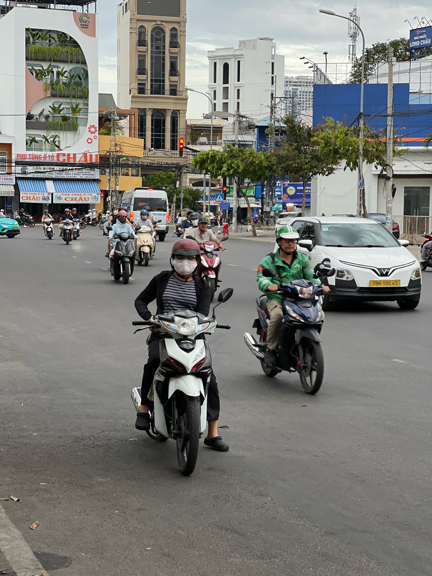 The image captures a street scene in Nha Trang, Vietnam, featuring a busy intersection with numerous motorcycles and a few cars. In the foreground, a person wearing a mask and a striped shirt rides a white and red motorcycle. To their right, another person in a green jacket and helmet rides a dark blue motorcycle. A white VinFast car with license plate 79H-100.45 is visible behind them. In the background, several other motorcycles, a white van, and various buildings of different architectural styles are present. The sky is overcast. The street is paved with asphalt, and traffic signals are visible. The buildings display signage in Vietnamese, indicating commercial establishments.