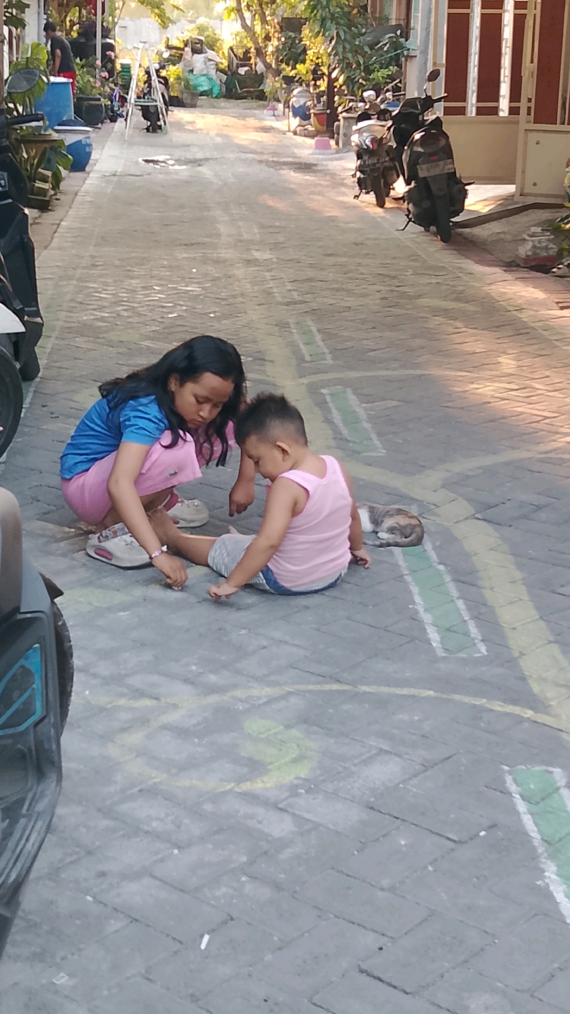 In Banjarsari, Indonesia, two children are on a paved path. A young girl with dark hair, wearing a blue shirt and pink shorts, is kneeling and appears to be tending to the younger boy. The boy, wearing a pink tank top and gray shorts, is seated on the ground. To the right of the boy, a calico cat is resting. The environment suggests an outdoor residential area with parked motorcycles in the background and chalk markings on the path.