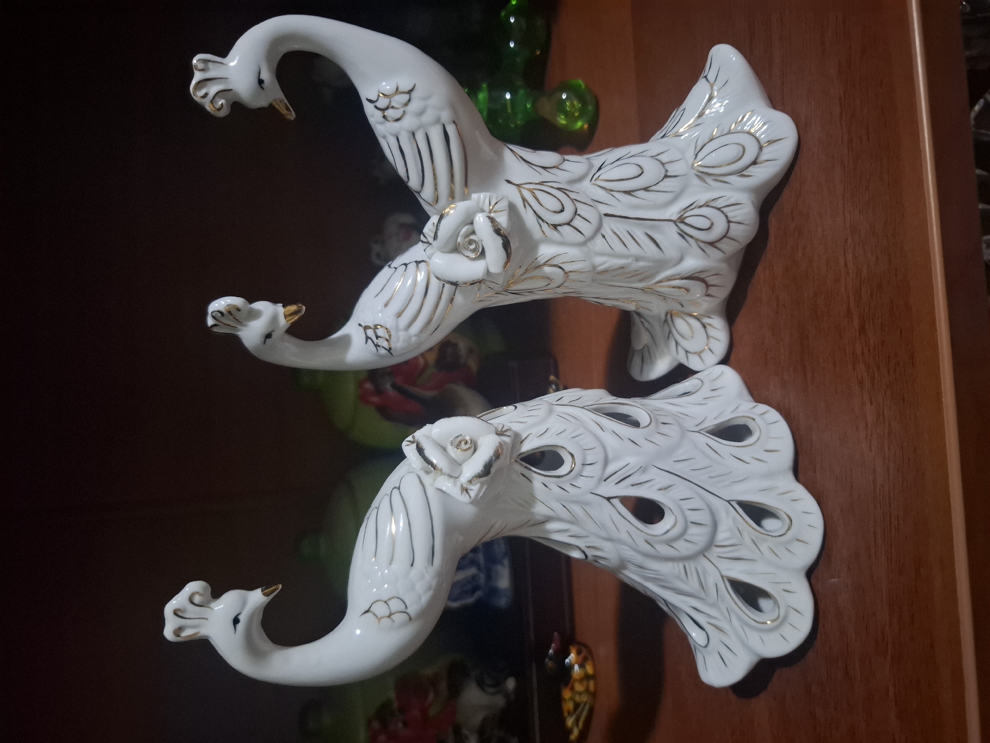Two white porcelain peacock figurines with gold accents are displayed on a wooden surface. The figurines are positioned facing each other and have intricate detailing, including stylized feathers and rose embellishments at the base of their necks. The background is dark and out of focus, suggesting they are on a shelf or table in a room. There is no indication of the city of Almaty, Kazakhstan.