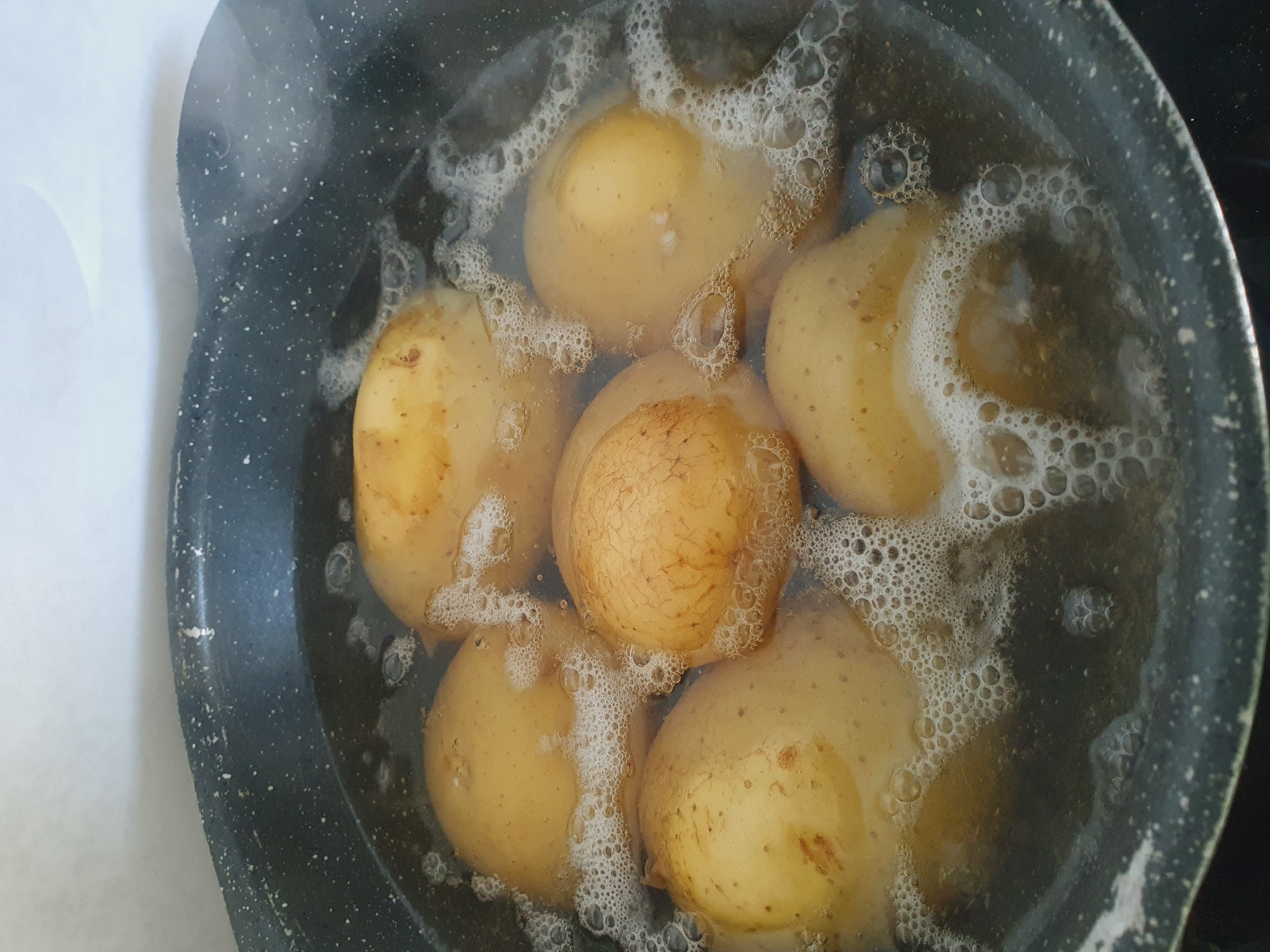A close-up, overhead shot shows several whole, unpeeled potatoes boiling in a dark gray pot. The potatoes are a light brown to yellow color, with some visible blemishes and lines on their skins. The water is cloudy and filled with small bubbles, indicating that it is actively boiling. The rim of the pot is visible, showing a speckled pattern with some wear. The background is a blurred, light-colored surface. The image appears to be taken in a kitchen setting.