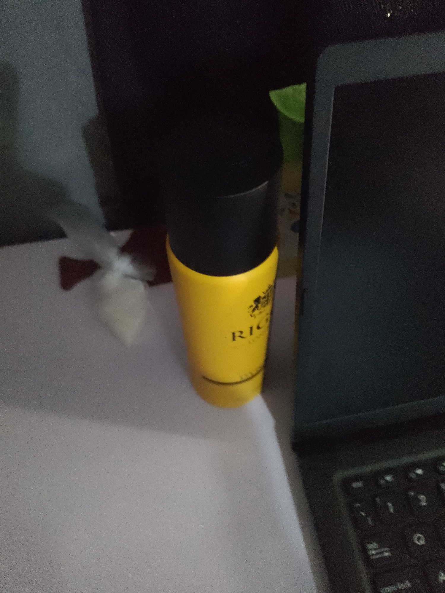 The image shows a close-up of a yellow cylindrical object with a black cap, possibly a spray can or a bottle, placed on a white surface. The object has some text on it, partially visible as "RIC". To the left of the object is a white crumpled paper-like item. In the background, a dark surface is visible, and to the right, a portion of a dark laptop screen and keyboard is in view. The lighting is dim, and the image has a grainy texture, suggesting it was taken in low light conditions. The scene appears to be indoors, likely on a desk or table. There are no people visible in the image, and no specific geographical landmarks or context are provided to determine its location within Jauro Yinu, Nigeria.