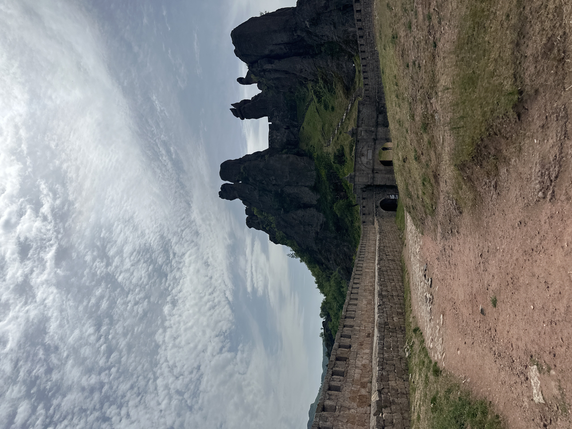 The image shows a landscape in Belogradchik, Bulgaria, featuring a distinctive rock formation adjacent to a stone wall, likely part of the Belogradchik Fortress. The rock formations are dark and jagged, contrasting with the lighter sky filled with textured clouds. The stone wall extends along the right side of the image, with several archways visible. The ground on the right side of the wall consists of reddish-brown dirt and sparse vegetation.