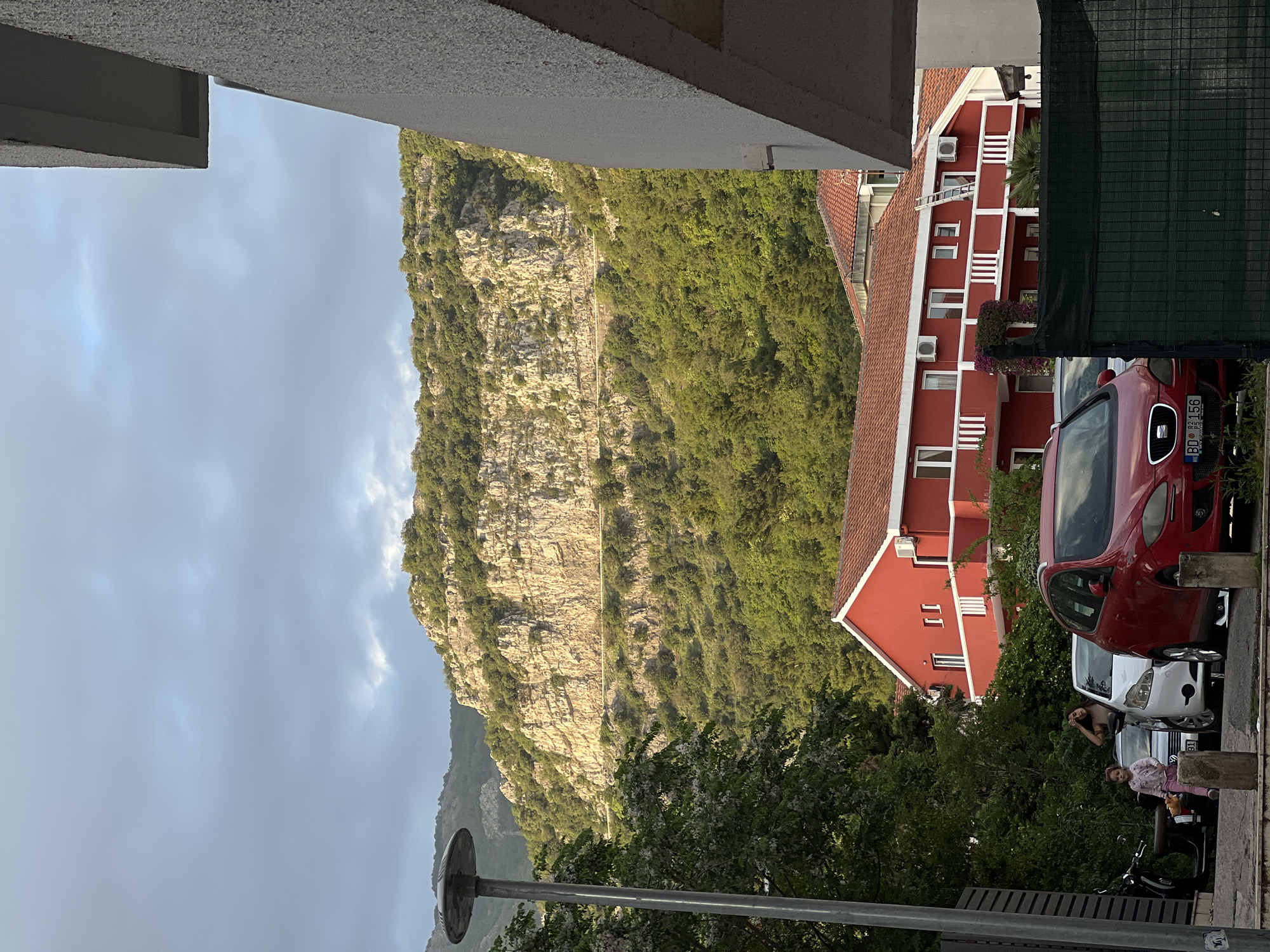 The scene is taken in Budva, Montenegro, showing a view dominated by a large, rocky mountain covered with green vegetation. The sky is overcast. A red building with white accents is visible to the right, and a portion of another building is seen above. Several parked cars are to the right, including a red car and a white car. In the foreground, there is a street lamp, some trees, and what appear to be people standing near the parked cars. A bench is seen near the street lamp.