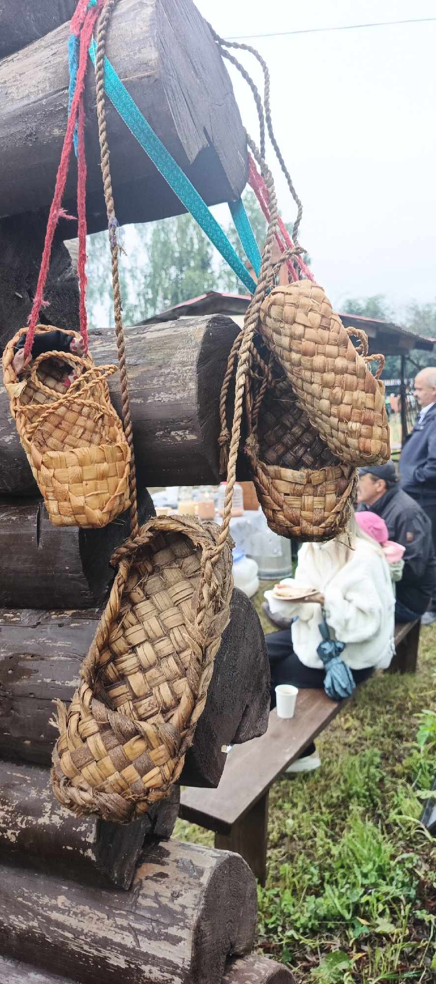 The image depicts an outdoor scene in the village of Starye Bygi, Russia, possibly at a market or cultural event. The foreground is dominated by several traditional Russian bast shoes (lapti) hanging from wooden beams by ropes and colorful ribbons. These shoes are woven from natural materials, likely bark or straw, and vary in size and condition.
Behind the shoes, a wooden bench is visible, occupied by several people. A woman in a white jacket is seated, holding a plate with food. To her right, another person is partially visible. In the background, other individuals are seen, including a man in a dark jacket. The setting appears to be rustic, with wooden structures and greenery. The sky is overcast, suggesting cloudy weather. Jars or containers are visible on a table in the mid-ground. The overall impression is of a casual gathering in a rural or historical setting.