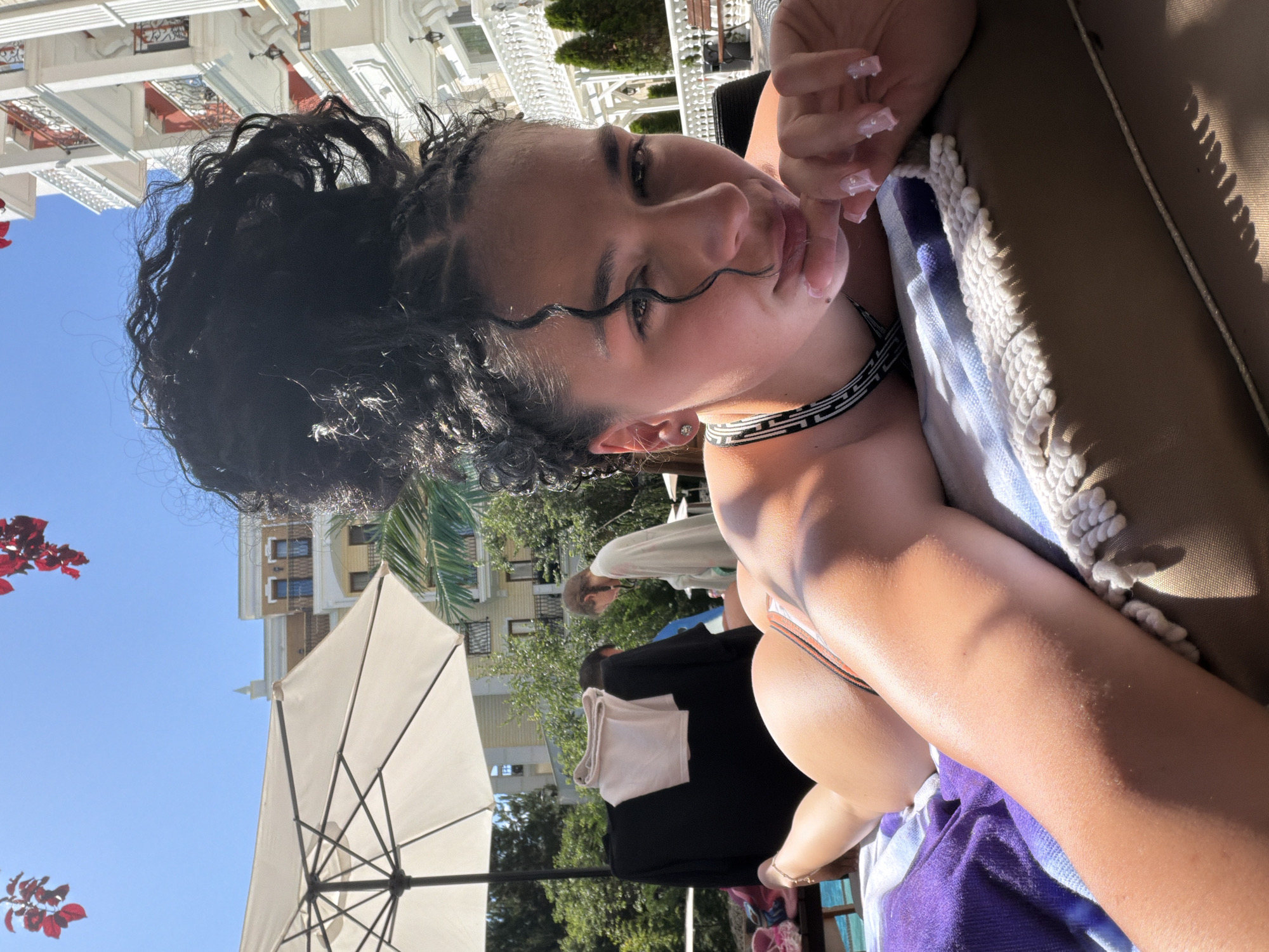 A young woman with dark, braided hair pulled into a high bun is sunbathing outdoors on a lounge chair. She is wearing a bikini and her body is partially visible. Her face is turned towards the camera, with one hand resting on her chin. In the background, there is a white umbrella, trees, a building with white balconies, and a clear blue sky. There are also other people in the background, one of whom is wearing a light-colored robe. The setting appears to be a hotel or resort area, likely in the city of Nesebar, Bulgaria, given the architectural style of the building.