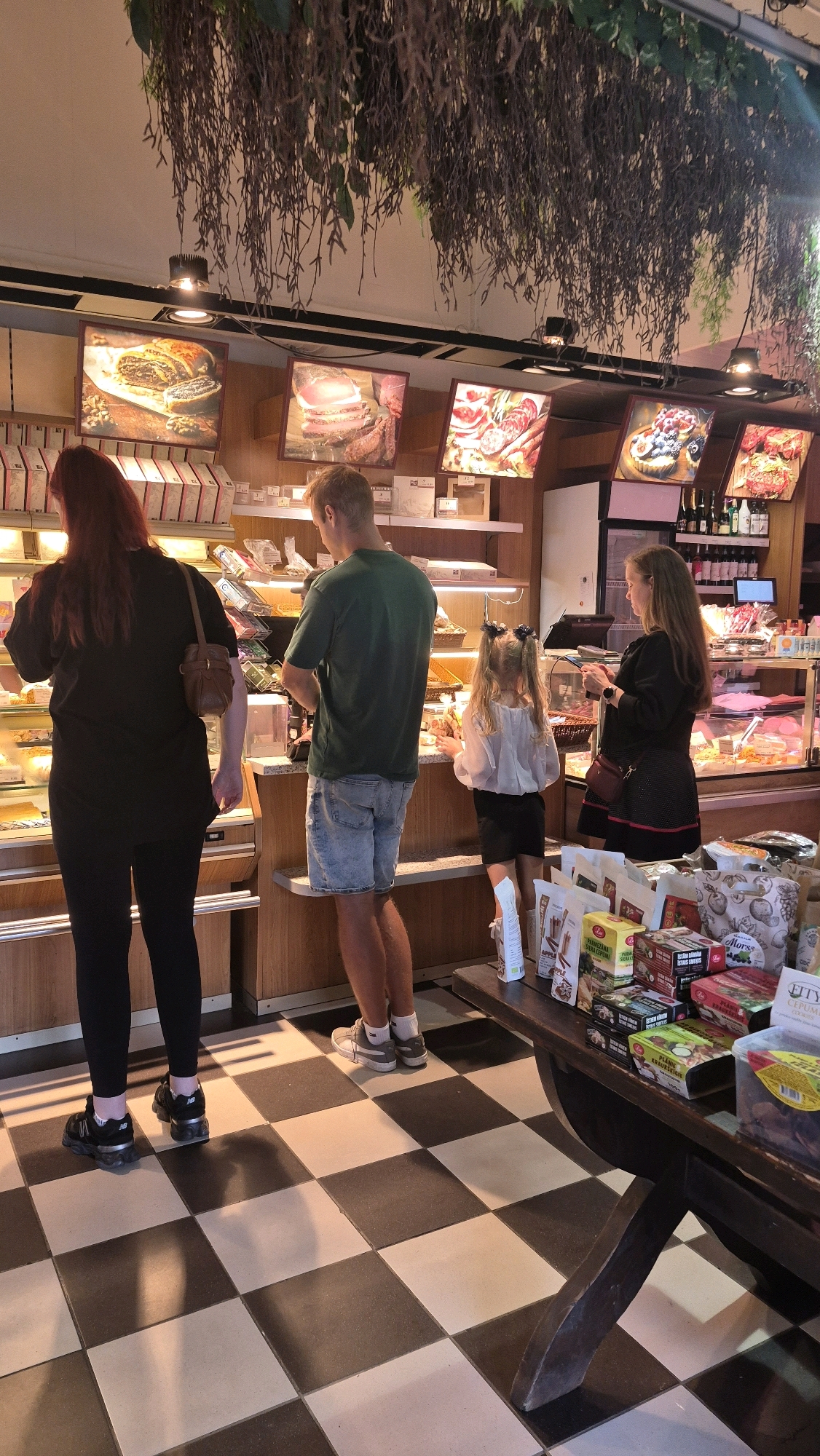 In Jēkabpils, Latvia, inside a retail establishment, possibly a bakery or deli, four individuals are observed. A woman with long red hair, wearing a black t-shirt and black leggings, stands on the left, facing a display case. To her right, a man in a green t-shirt and denim shorts is looking into a display. A young girl with blonde hair in pigtails, wearing a white blouse and black skirt, stands between the man and another woman. The second woman, with long brown hair, is dressed in a black dress and is holding a mobile phone. The environment features checkered flooring, wooden shelves stocked with various items, and framed pictures of food products displayed on the wall. The scene appears to depict customers browsing or making purchases.