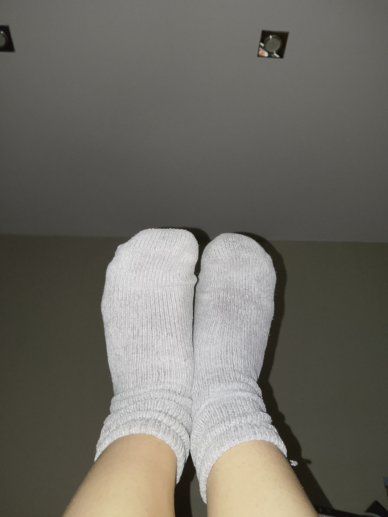 This is a close-up, waist-up photograph of two bare legs wearing white, ribbed socks. The legs are angled upwards and appear to be positioned on a surface or held up. The background is a plain, dimly lit wall with two circular, recessed light fixtures visible in the upper portion of the image. The overall lighting suggests an indoor environment, likely a residential setting. There are no people or other significant objects in the frame besides the legs and socks. The specific location within Ostróda, Poland, cannot be determined from the visual information provided in the image.