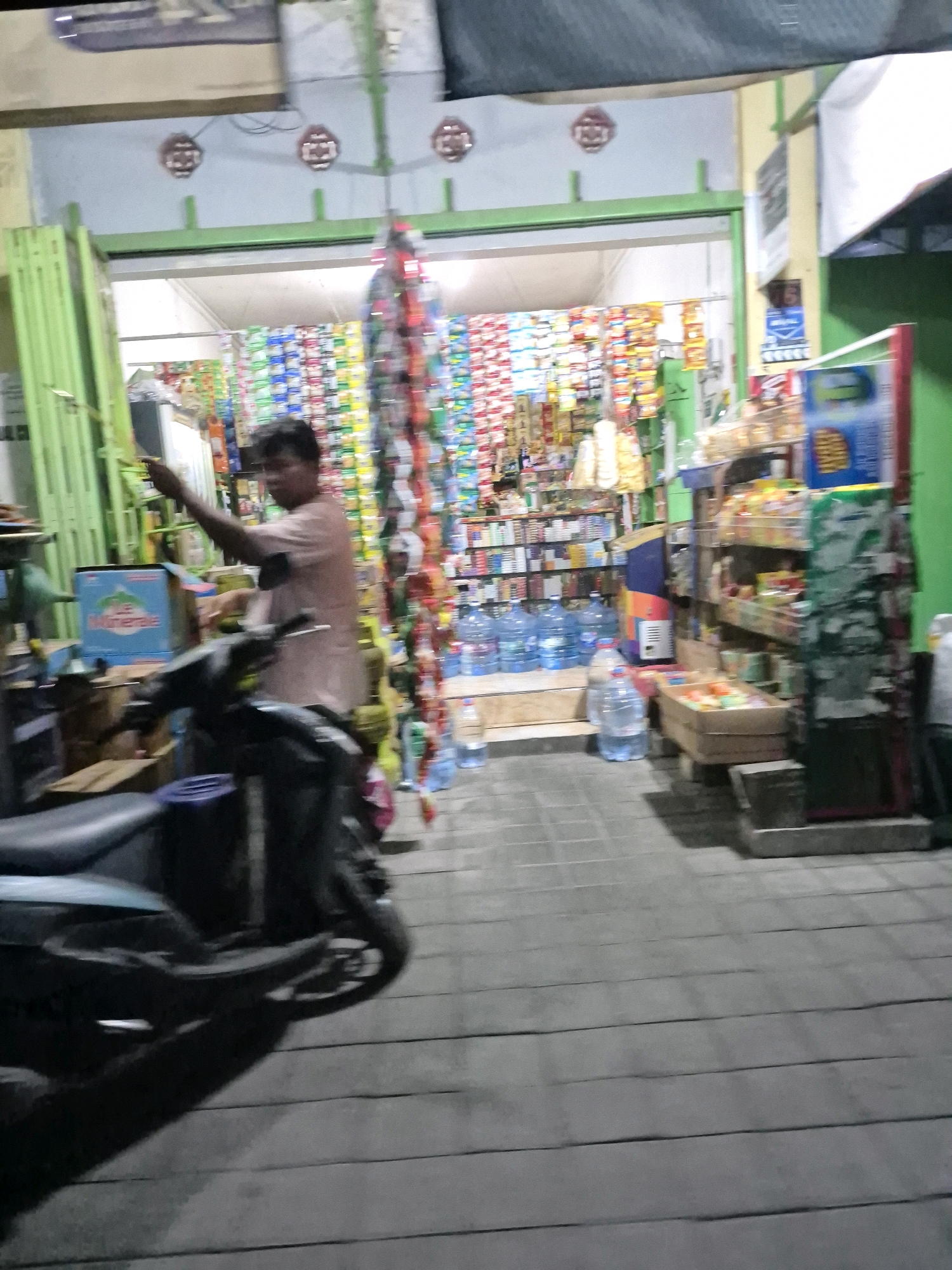 A man is standing in front of a store in Pabelan, Indonesia. He is wearing a light brown t-shirt and appears to be pointing to something. In the foreground, a dark-colored motorcycle is parked. The store is stocked with various items, including bottles of water and snacks, visible on shelves and displays. The store has a green metal gate that is partially open, revealing the interior. The ground in front of the store is paved with large, rectangular tiles. The overall scene suggests a small convenience store or warung operating at night.
