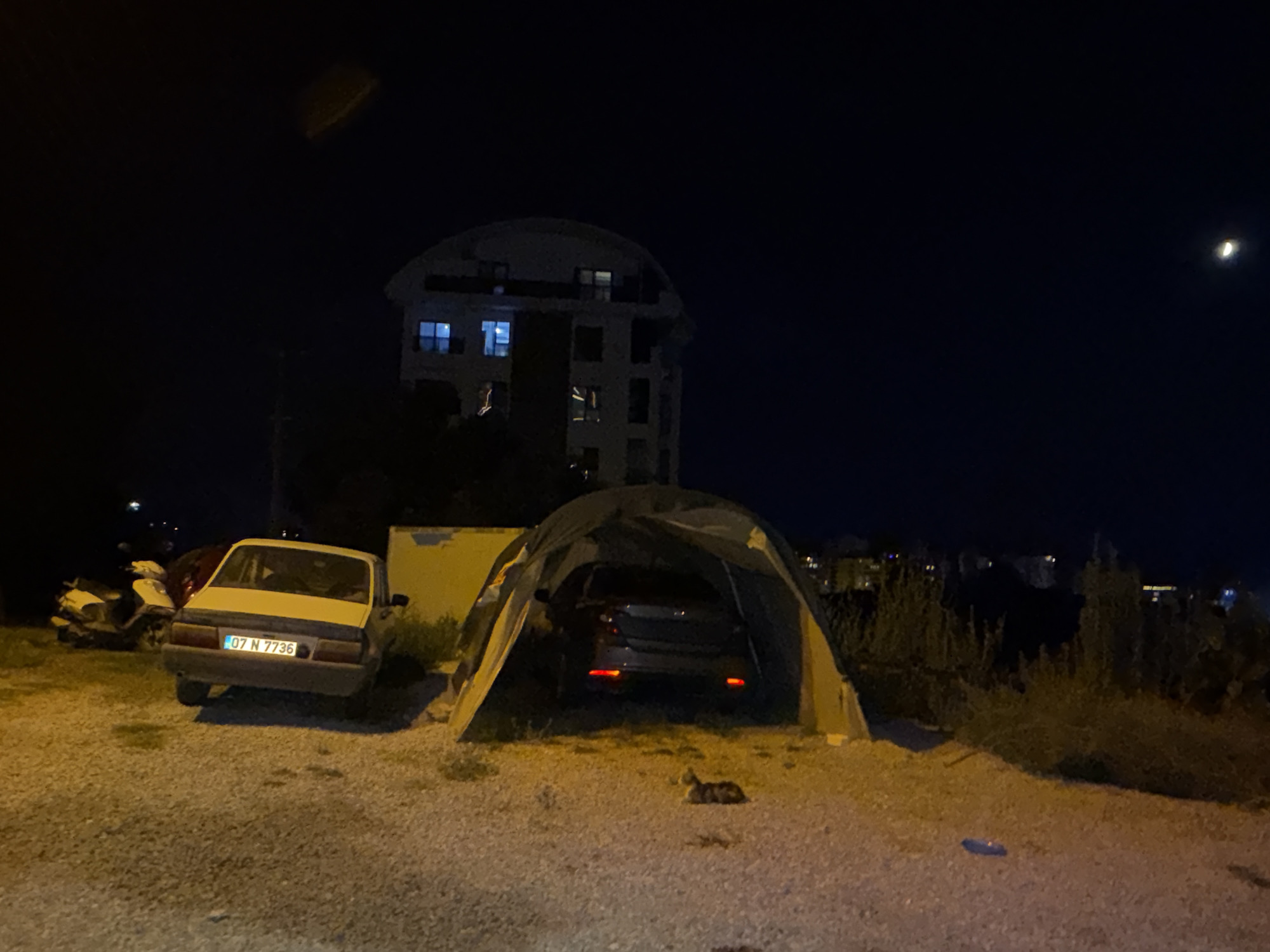 The image depicts an outdoor scene at night in Alanya, Turkey. In the foreground, a gravel area is visible with a yellow and grey tent set up. A dark-colored car is parked inside the tent. To the left of the tent, a light-colored car with a license plate reading "07 N 7736" is parked. A white scooter is also parked to the left of the car. In the background, a multi-story building with illuminated windows is situated against a dark night sky. A crescent moon is visible in the upper right corner of the sky. There is a small tabby cat lying on the gravel in front of the tent. The scene appears to be a temporary encampment or storage area in a semi-urban or rural setting.