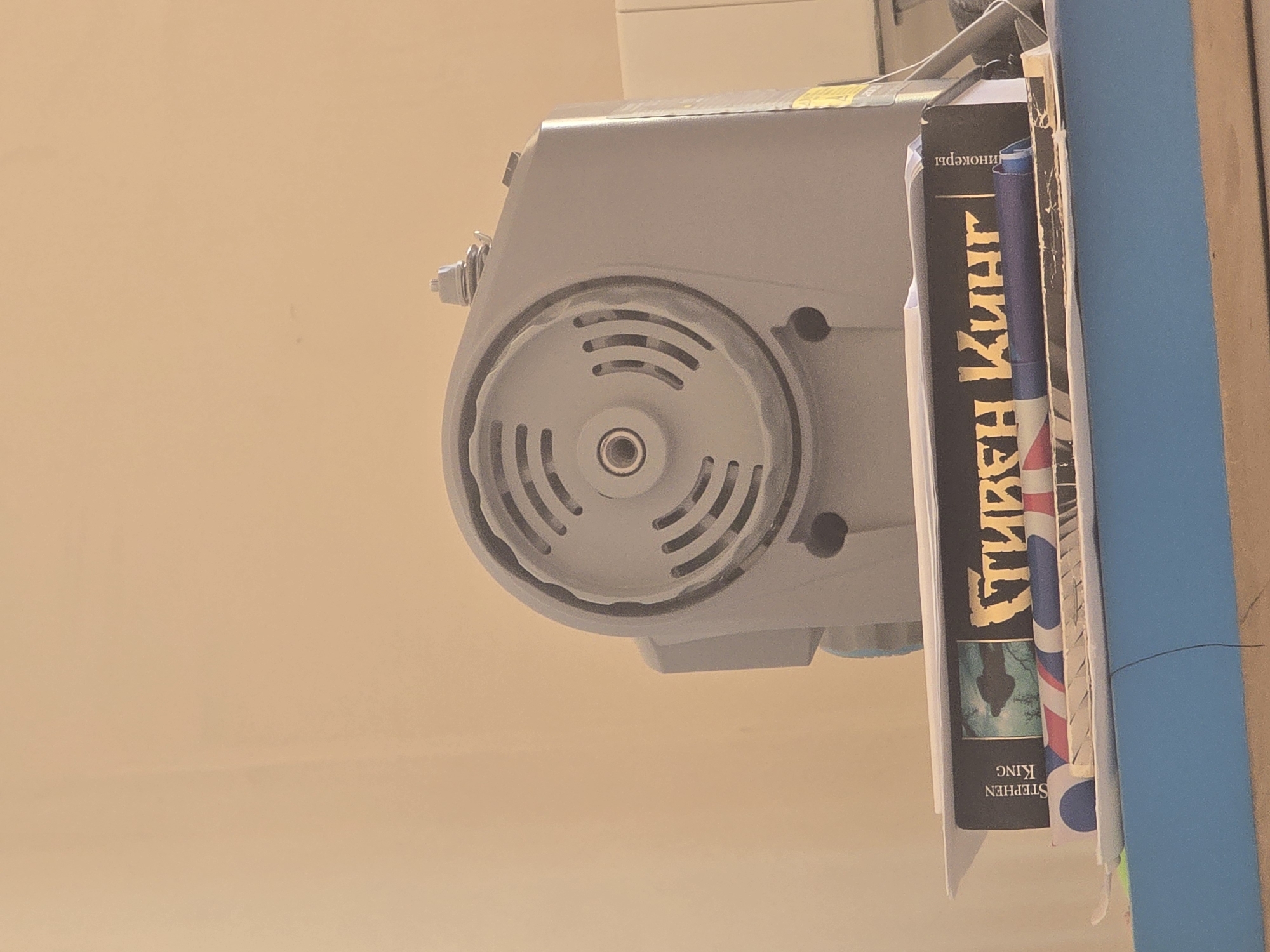 The image shows a close-up of a sewing machine, a book, and other items on a shelf. The sewing machine is gray and has a circular vent on the top. The book is black with gold lettering and appears to be in Russian. The spine of the book has the name "STEPHEN KING" printed vertically. A thin black wire is visible in the foreground. The background is a plain, light-colored wall. The scene is indoors, on a shelf, likely in a domestic setting. There is no information to determine the specific location in the city of Avtury, Russia.