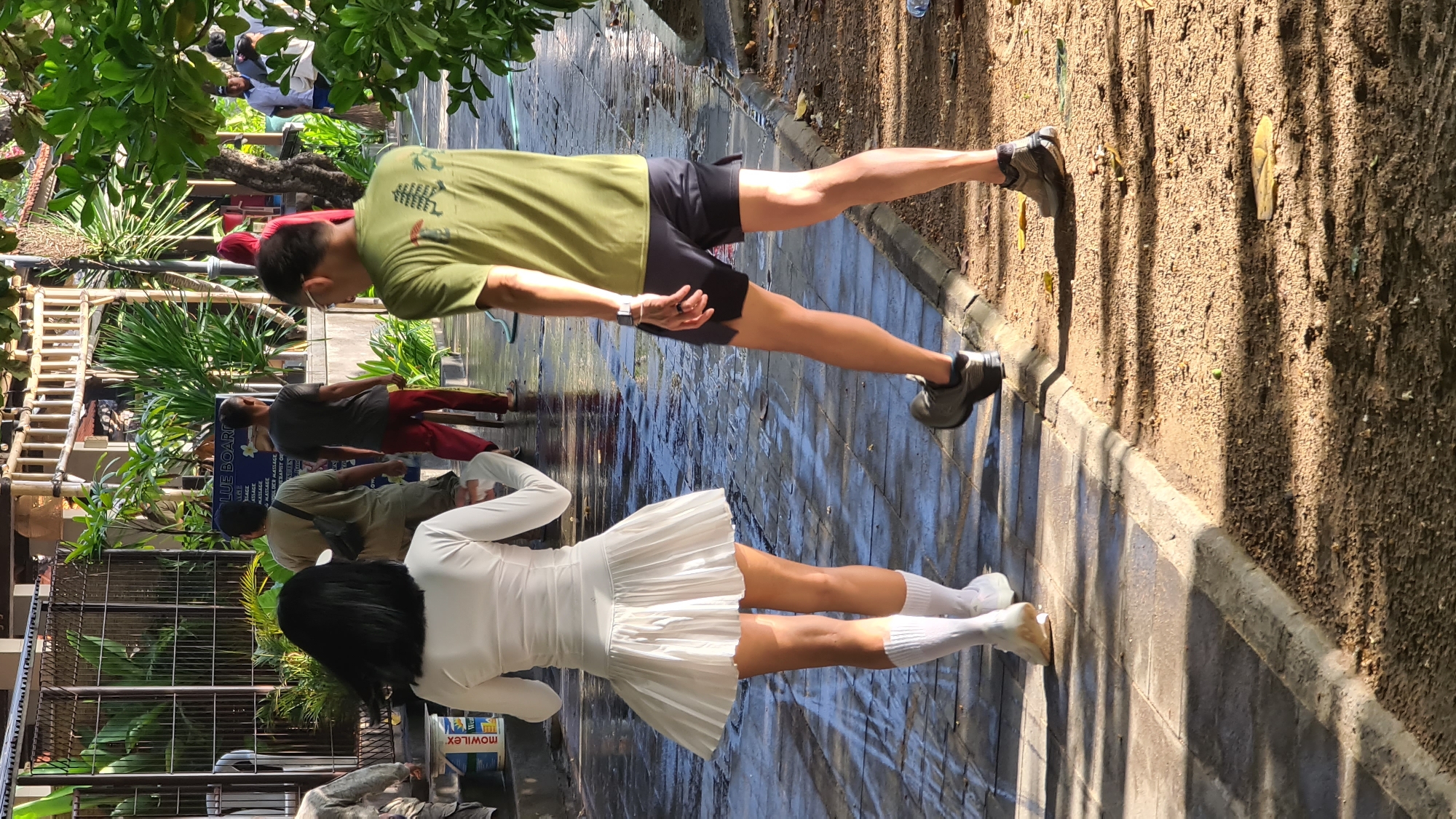 In Denpasar, Indonesia, on a paved walkway with some areas of dirt and vegetation, two individuals are walking away from the camera. The person on the left is a female wearing a white long-sleeved top and a white pleated skirt, with white socks and white shoes. She has black hair. The person on the right is a male wearing a green t-shirt and dark shorts, with grey athletic shoes. They appear to be walking in the same direction. In the background, there are other individuals, some carrying bags, and a sign that reads "BLUE BOARD." The walkway is wet, suggesting it may have recently rained or been cleaned. Sunlight casts shadows on the ground, indicating it is daytime.