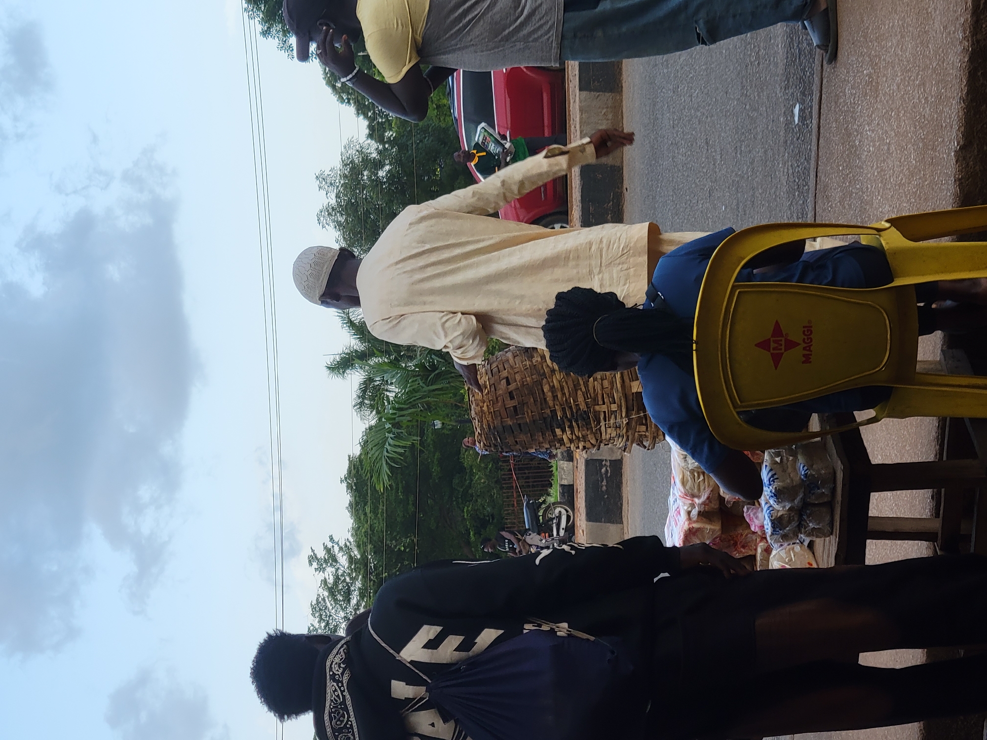 The image captures a street scene in Jos, Nigeria, likely a market area due to the presence of vendors and goods. In the foreground, a person wearing a black hoodie and a blue backpack stands facing away from the camera. To their right, a man in a light-colored garment and a white cap is also facing away, carrying a large woven basket. Further to the right, another individual in a yellow and grey shirt, wearing a dark baseball cap, is seen in profile with their hand to their ear, possibly on a phone. In the middle ground, a red vehicle is parked on the road, and a man in a green shirt is holding what appears to be a stack of items. A motorcycle is visible behind the man with the basket. The background shows lush green trees and foliage, suggesting an outdoor setting within the city. In the immediate foreground, a table is laden with packaged goods, likely bread, and a yellow plastic chair with a "MAGGI" logo is occupied by a person wearing a blue shirt. The overall atmosphere is that of an active, everyday street life in Jos.