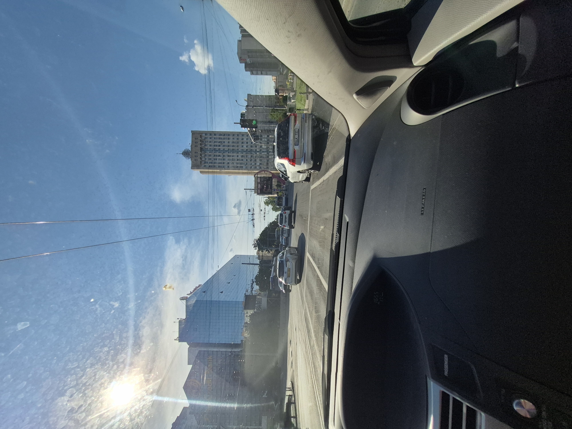 The image is taken from inside a car, looking forward through the windshield. The car is driving on a multi-lane road in a city. On the left side of the road, there are several modern glass-fronted buildings. One of these buildings appears to be an office building with a sign that reads "OTP Bank". Further down the road on the right, a tall, rectangular building with many windows is visible. There are also several other cars on the road, some in front of and some beside the car from which the photo is taken. The sky is blue with some scattered clouds, and the sun is shining brightly, causing glare on the windshield. The dashboard of the car is visible in the foreground. The scene appears to be a typical urban environment during the daytime. Based on the architecture and the presence of a green traffic light, the location is likely Chișinău, Moldova, specifically on or near Ștefan cel Mare și Sfânt Boulevard, a major artery in the city, near the intersection with Bucuriei Street, where a prominent landmark building of that architectural style can be found.