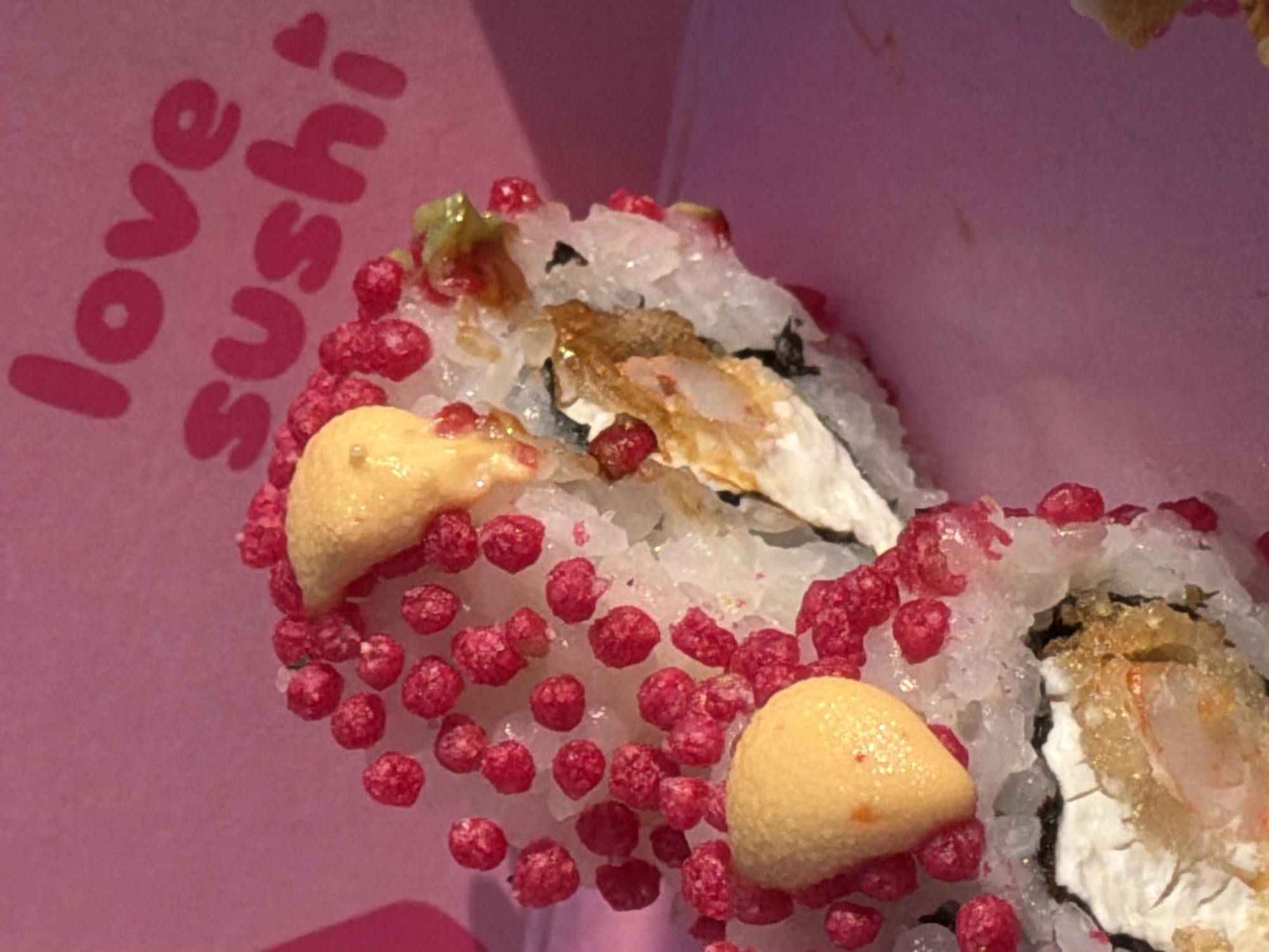 The image shows a close-up view of sushi rolls arranged in a pink box. The sushi appears to be of the California roll variety, with rice, seaweed, and fillings visible. The rolls are decorated with small, red, spherical toppings that resemble roe or a similar type of garnish. There are also dollops of a yellowish sauce, possibly mayonnaise-based, on some of the sushi pieces. The background of the image is a soft pink, which is consistent with the color of the box. The text "love sushi" is prominently displayed in a playful font on the pink surface of the box, with a small heart above the "i". The lighting suggests an indoor setting, possibly a restaurant or a takeout container. No people are visible in the image, and the focus is entirely on the prepared food. The overall impression is one of colorful and appealing sushi, presented in a branded container.