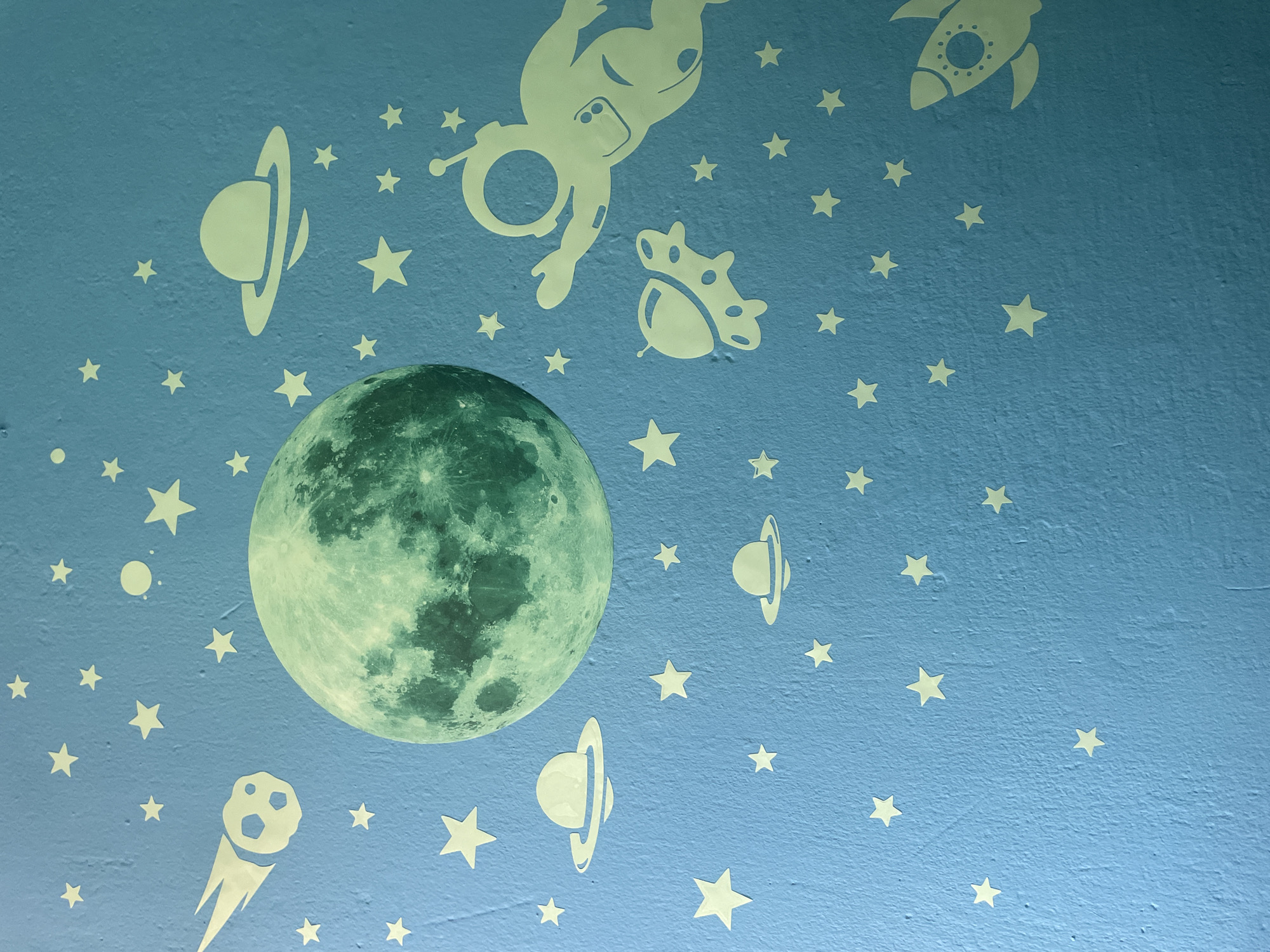 The image shows a blue, textured wall decorated with glow-in-the-dark stickers of space-themed objects. The primary subject is a large, green-toned sticker of the moon, positioned on the left side of the frame. Scattered around the moon are numerous small, white star stickers, some appearing to glow.
Several other white stickers depict various celestial bodies and spacecraft. These include:
- A stylized rocket ship at the top right.
- An astronaut in a floating pose, near the top center.
- A circular object with a ring, resembling a planet like Saturn, appears multiple times.
- A comet with a trailing tail, positioned on the bottom left.
- A UFO-like object near the astronaut.
- A small, solid circle, possibly representing another planet or celestial body.
The scene is clearly indoors, on a wall, likely in a child's room or playroom, given the playful space theme. There is no indication of the time of day or weather, as it's an indoor setting. The overall impression is one of whimsical fantasy and exploration of space. The glow-in-the-dark aspect of the stickers suggests the image may have been taken in low light conditions to highlight their luminescence. No visible text is present in the image. The location context provided (Kołobrzeg, Poland) does not offer any visual cues within the image itself.