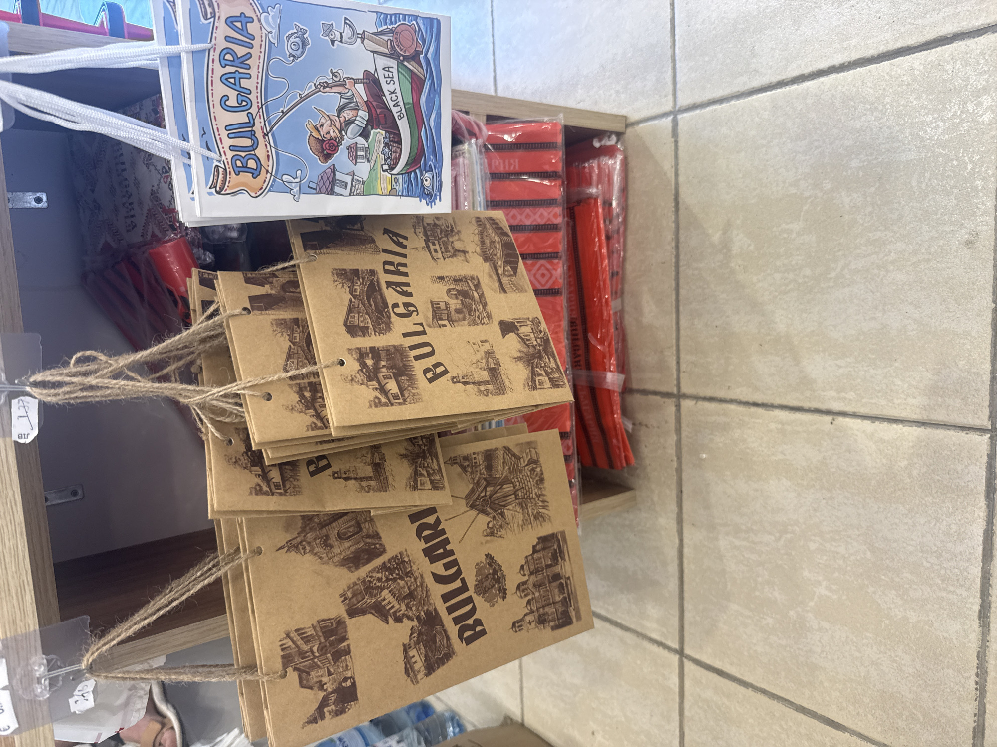 This image shows a close-up, angled view of a retail display of souvenirs. The primary objects are several stacks of brown paper items, likely postcards or small bags, each printed with images of Bulgarian landmarks and the word "BULGARIA." These are suspended by twine from clear plastic clips attached to a wooden shelf.
To the left, a postcard featuring a cartoon illustration of a person fishing from a boat labeled "BLACK SEA" and "BULGARIA" is also visible, attached by white string. Behind these items, a shelf holds several red items wrapped in plastic, possibly textiles or other souvenirs, with some Cyrillic text visible on the packaging.
The setting appears to be an indoor shop, suggested by the wooden shelf and the tiled floor in the background. The lighting is bright, indicating daytime. There are no people directly visible in the frame, but a person's head and shoulder are partially visible at the bottom left, suggesting they are browsing or working in the vicinity. The overall impression is a display of tourist mementos. Visible text includes "BULGARIA," "BLACK SEA," and some Cyrillic characters. The location context is Varna, Bulgaria.