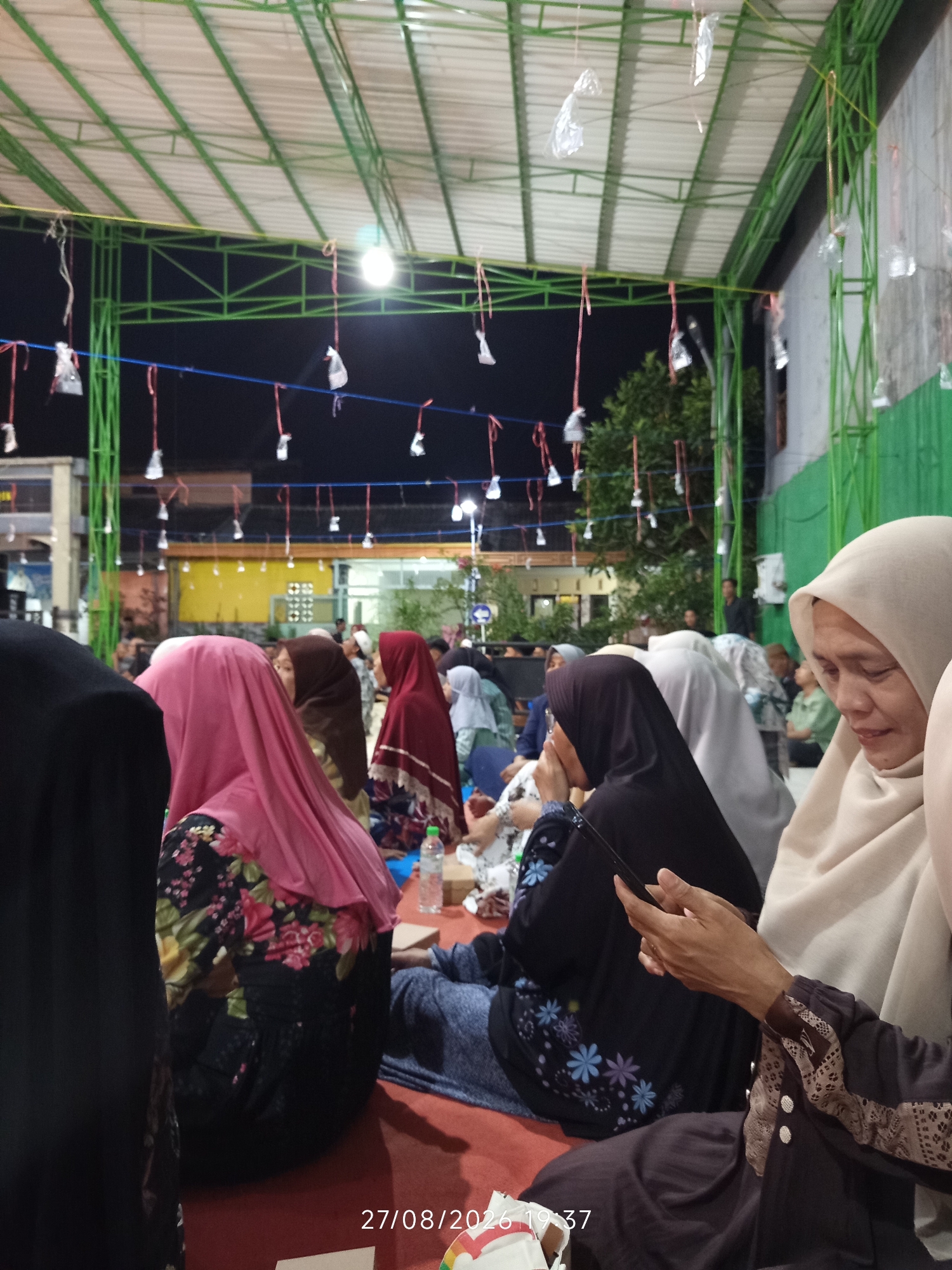 The image shows a group of people, mostly women wearing hijabs, gathered outdoors under a green metal-framed roof at night. The roof is translucent white and supported by green struts. Numerous small, white objects wrapped in plastic are hanging from thin red cords suspended from the roof structure, suggesting a decorative or ceremonial purpose. The background features a dark night sky, some trees, and buildings with yellow and orange lighting. In the foreground, several individuals are seated on a red carpet. One woman in a light brown hijab is in focus, looking down and to the left, with her hands resting on her lap. Another woman wearing a black hijab is also in the foreground, partially obscured. The overall scene suggests a community gathering or event taking place in an outdoor, semi-enclosed space. The timestamp "27/08/2026 19:37" is visible in the bottom right corner, indicating the date and time the photo was taken.