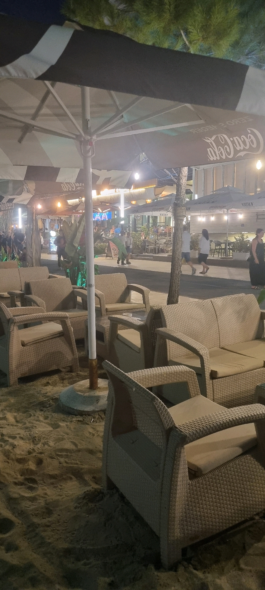 An outdoor seating area is depicted at night on a sandy surface, likely a beach or promenade, in Bashkia Durrës, Albania. The scene is illuminated by artificial lighting, casting shadows and highlighting the textures of the environment. In the foreground, several wicker armchairs and a sofa are arranged around a glass-topped table. A large, striped umbrella, partially covering the furniture, features a Coca-Cola logo printed on its underside. Behind the seating area, a pathway is visible, with people walking and standing. Several structures, including cafes or shops with awnings and tables, line the pathway. One awning displays the text "VATAN". The overall ambiance suggests a casual, nighttime establishment.