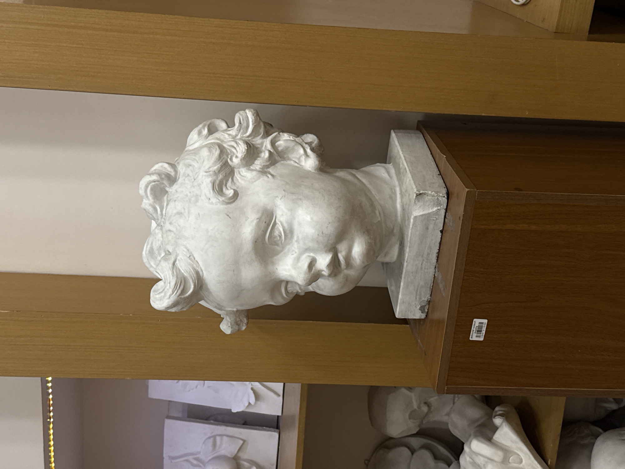 A white plaster bust of a child's head sits atop a dark brown wooden shelf within a retail or display setting. The bust, featuring detailed curls, is placed slightly off-center towards the left. The shelf is surrounded by other plaster objects on a lower shelf, partially visible. The bust is in Yerevan, Armenia.