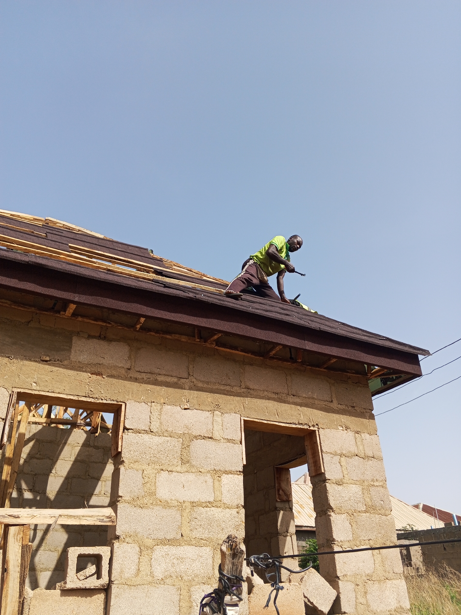 A construction worker is installing roofing shingles on a cinder block building in Unknown, Unknown. The building has openings framed in wood for doors and windows. Some roofing shingles have already been installed on the roof, and the wooden framework beneath is visible in areas where shingles are missing. The sky is clear and blue. Power lines are visible in the background. Debris, including cinder blocks and construction material, are visible in the foreground.
