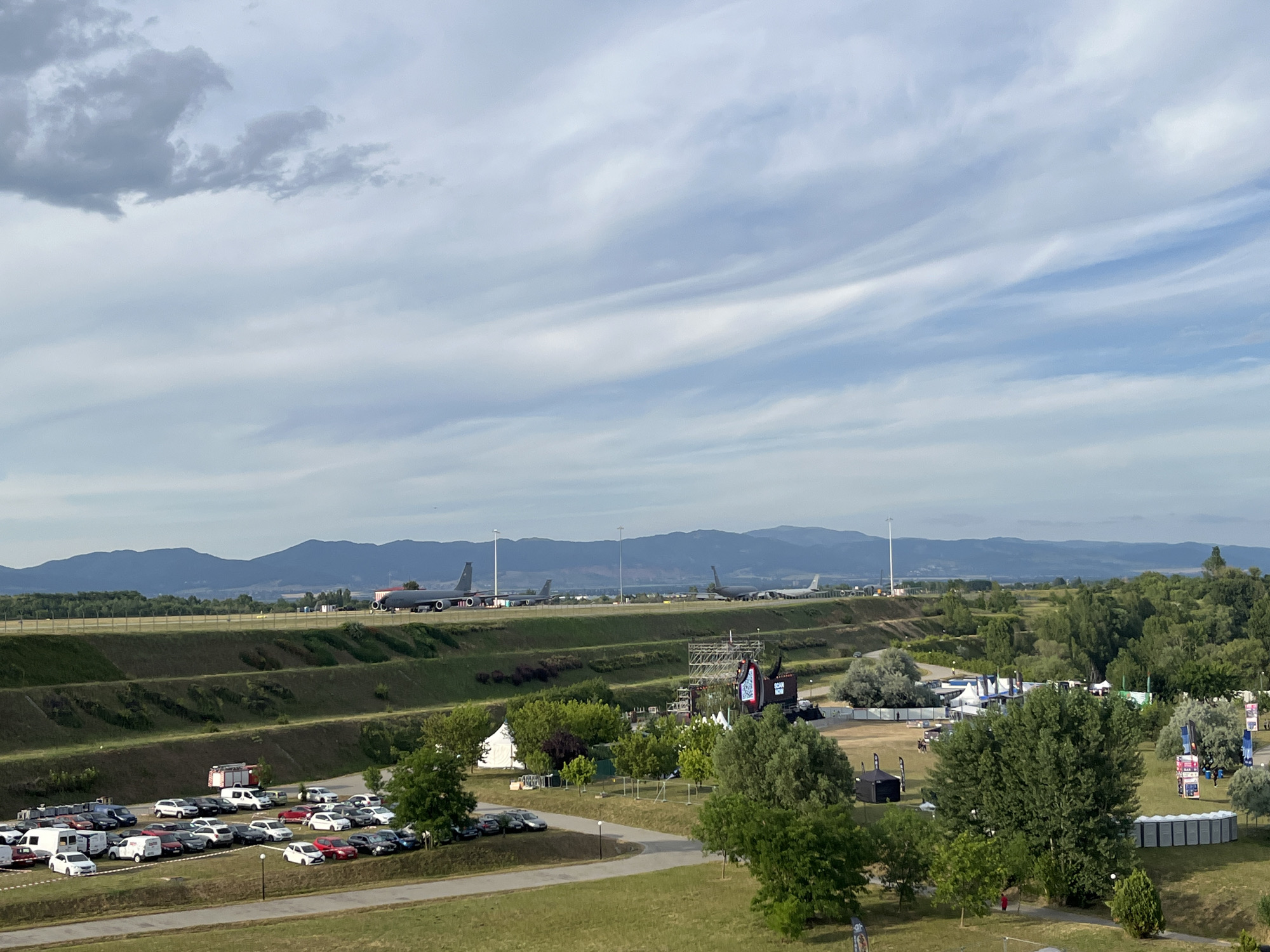 The image shows an outdoor event possibly an airshow or festival, set against a backdrop of rolling hills and mountains under a partly cloudy sky. In the foreground, a large parking lot is filled with numerous cars and some vans. A fire truck is parked near the edge of the lot.
Further back, a series of tiered green slopes lead up to an elevated area where several large, dark-colored airplanes are parked on a tarmac. These appear to be military aircraft, possibly bombers or tankers, given their size and shape. The planes are spaced out along the tarmac.
In the middle ground, a festival or event setup is visible with temporary structures, tents, stages, and a large screen displaying "SCAN NOW". Trees and grassy areas surround these structures.
In the far background, a range of blue-hued mountains forms the horizon. The overall setting suggests an open area, likely an airfield or a large park, near an urban or semi-urban area, given the presence of the airplanes and the event infrastructure. The event appears to be taking place during daylight hours, possibly late afternoon, judging by the light and shadow. The location is the Sofia Airport area, specifically the Aviation Museum or surrounding grounds, which occasionally host such events.