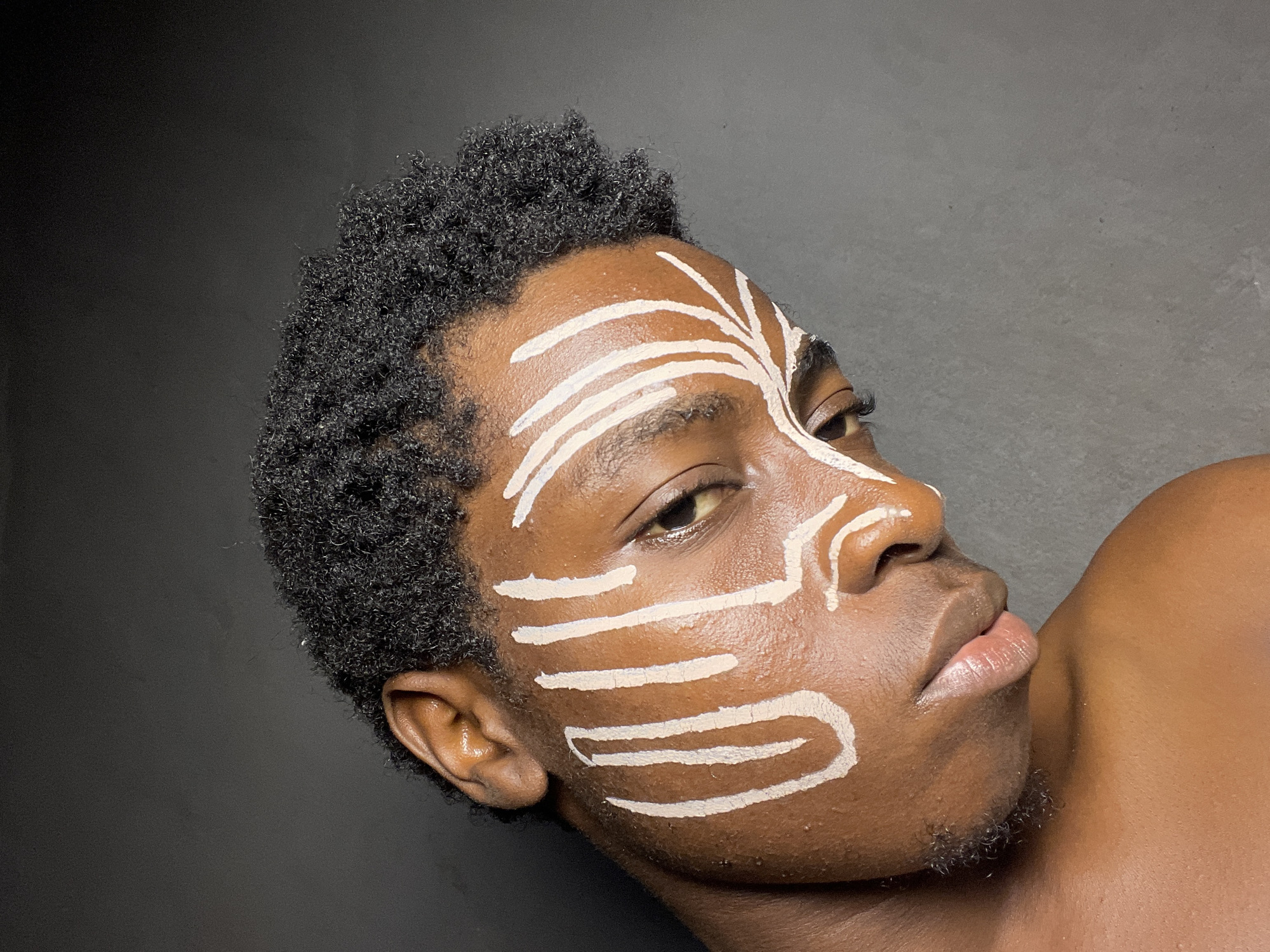 The image shows a close-up, head and shoulders shot of a young Black person against a dark gray background. The subject's skin is a rich brown tone, and they have dark, curly hair. White face paint is applied in a tribal or artistic pattern on the left side of their face, extending from the forehead down to the cheekbone and around the eye. The paint consists of several bold, vertical strokes on the forehead and temple, with thinner, radiating lines fanning out towards the cheek. The subject is looking downwards and slightly to the right, with their head tilted. Their lips are slightly parted. The lighting emphasizes the contours of their face and the texture of the paint. No specific location in Nkpor, Nigeria is discernible from the image itself.