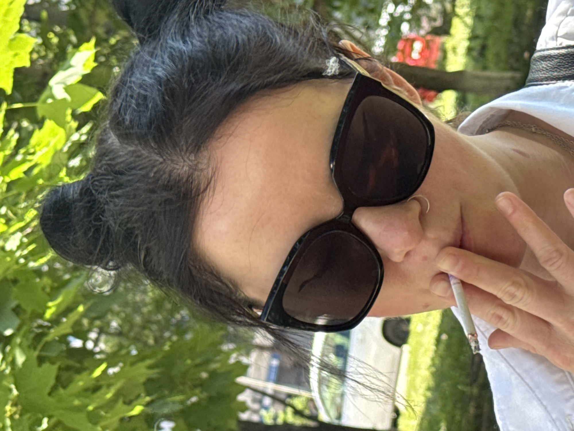A close-up, slightly angled shot shows a person from the shoulders up, outdoors on a sunny day. They have dark hair styled in two buns on top of their head, and are wearing large, dark sunglasses. The person is holding a lit cigarette in their right hand, with their index and middle fingers around it, and bringing it to their lips. They are wearing a white collared shirt and a silver chain necklace. A nose ring is visible. In the background, out of focus, are green leaves from trees, a white car, and hints of red structures. The overall environment appears to be a park or street with foliage.