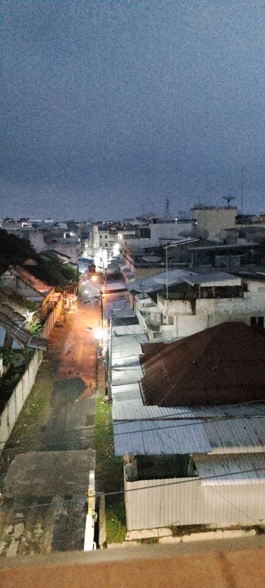 The image depicts an aerial view of a street in the City of Medan, Indonesia, at dusk or night. The street is paved and appears wet, with reflections from overhead lights. Buildings line both sides of the street, characterized by tiled roofs, some showing signs of age. The architecture is varied, with multi-story structures and single-story dwellings visible. Some buildings have balconies and windows. Streetlights illuminate parts of the scene, casting a warm glow. Vegetation, including grass and some trees, can be seen along the sides of the street and between buildings. The sky is overcast and a dark blue-gray. The overall impression is of a residential or commercial area in an urban environment.