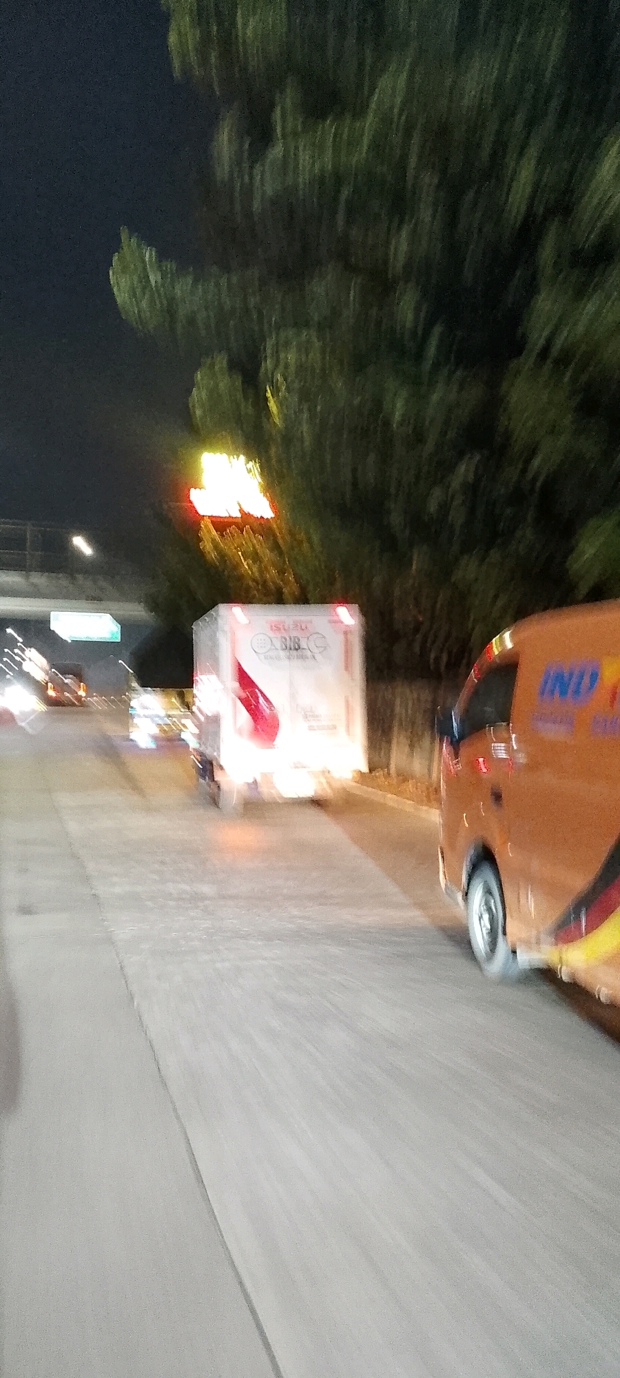The image depicts a night scene on a multi-lane road, likely an expressway or highway, in Depok, Indonesia. The perspective is from a vehicle in motion, indicated by the motion blur. In the foreground, on the right, is the rear side of an orange van with black and yellow striped accents. The van has visible text that appears to read "IND" in blue lettering on its side. To the left of the van, and slightly further down the road, is a white box truck. The truck has red taillights illuminated, and on its side, the word "ISUZU" is visible above a logo that includes the letters "BIB" and what appears to be a wrench. The truck also has a red graphic element. Further down the road, other vehicles are visible as streaks of light, suggesting ongoing traffic. Overhead, a lit sign with bright yellow and red lettering is visible, partially obscured by dark green foliage. The road surface is grey and appears to be asphalt, with lane markings faintly visible due to the motion blur. The environment suggests an urban or suburban setting with trees lining the side of the road. The overall impression is one of a busy nighttime commute or transit.