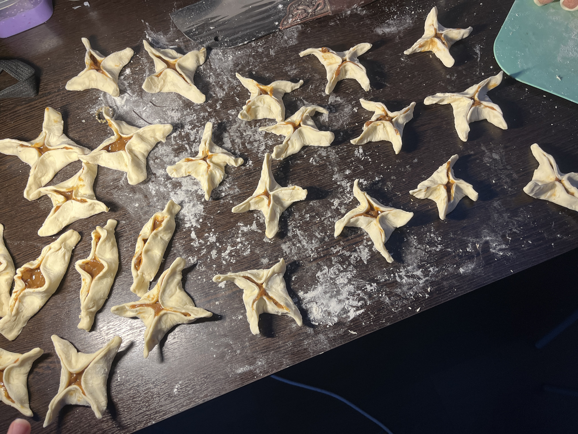 The image shows a dark brown wooden table with several uncooked pastries arranged on it. The pastries appear to be star-shaped with a caramel-colored filling in the center. There's a sprinkling of white flour on the table, suggesting the dough was prepared there. Visible items include a gray strap, a knife with an ornamental handle, and a light blue cutting board. The lighting appears to be natural. There is no indication of the location being Saint Petersburg, Russia, or any information about people.