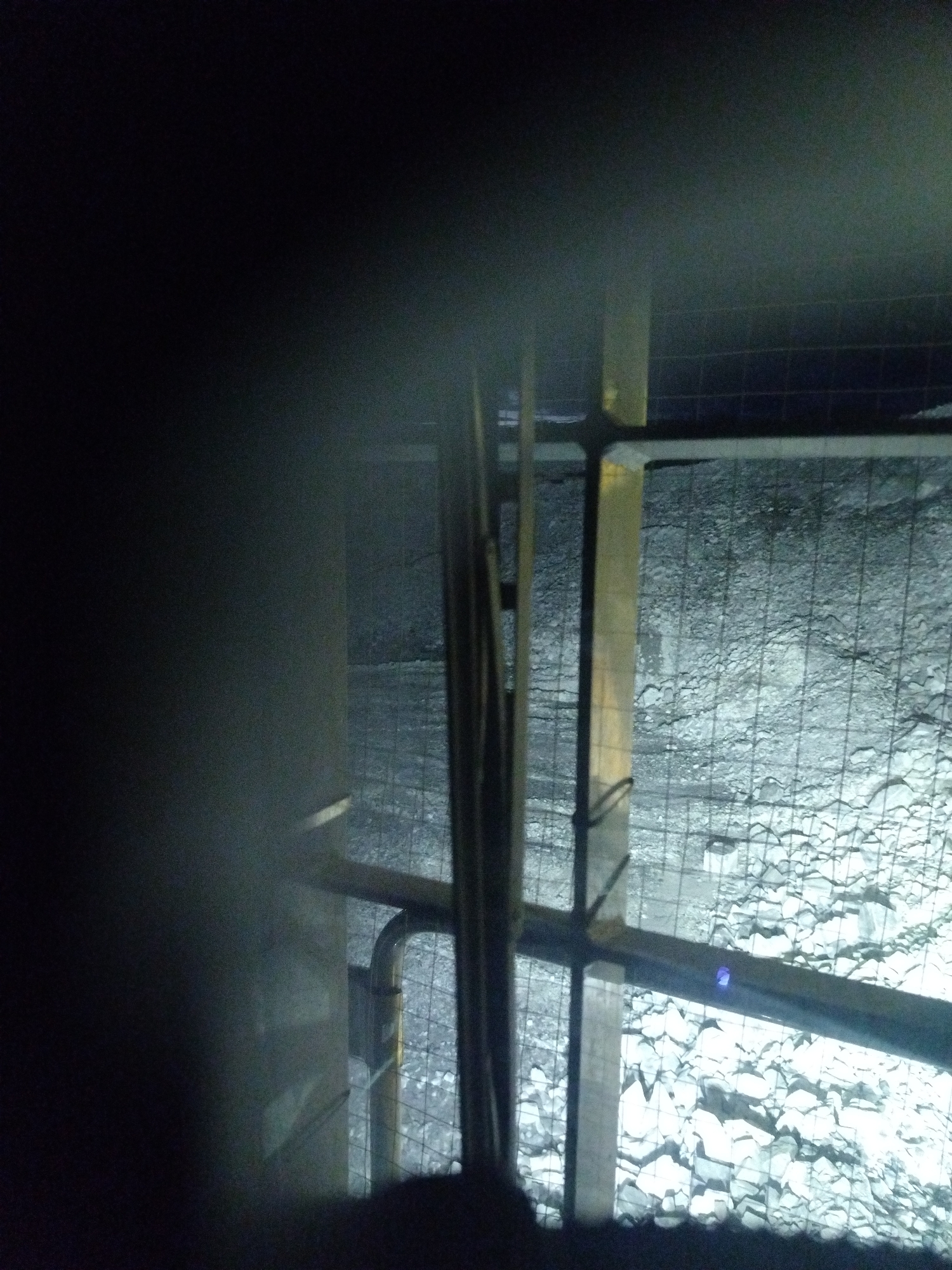 An image taken at night in an industrial or construction setting. The foreground is dominated by a dark, blurred area on the left, possibly a person or object obscuring the view. A metal structure, appearing to be scaffolding or part of a building, is visible in the mid-ground. Vertical and horizontal beams, some possibly painted yellow, form a frame. Within this frame, a wire mesh fence is apparent, separating the viewer from the background. The background itself consists of a rough, uneven surface covered in rocks and debris, illuminated by a cool, artificial light source that casts harsh shadows. There are no discernible people or specific actions taking place, suggesting a static or paused scene. The overall impression is one of a work in progress or an operational industrial area at night. The location in Naminga, Russia is not evident from the visual information provided in the image.