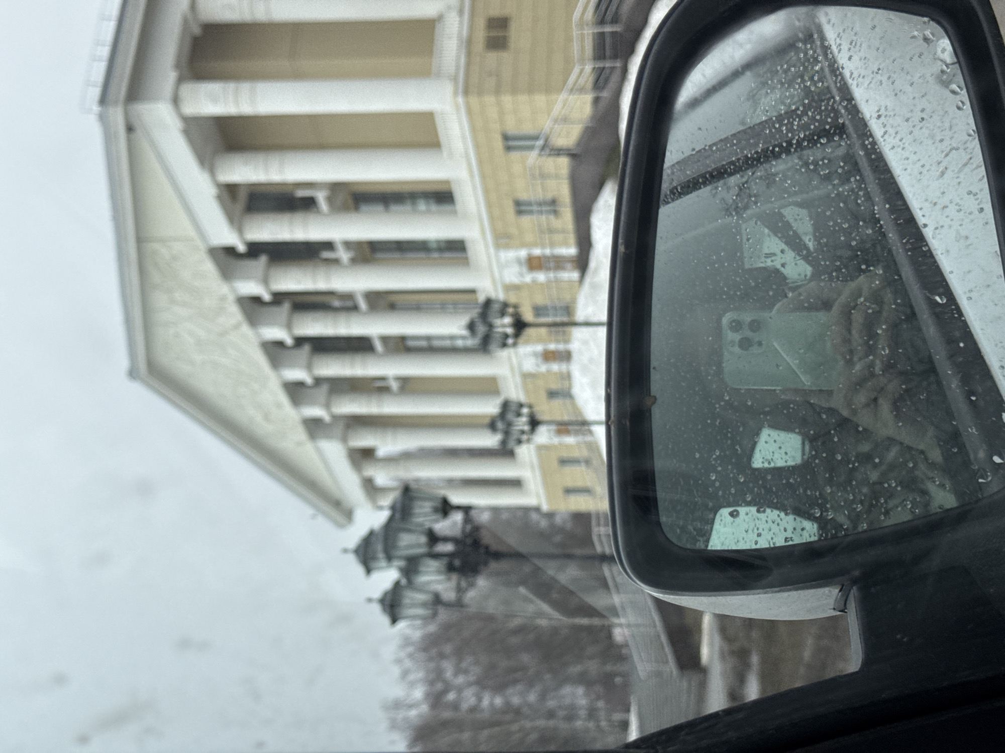 The image shows a view from inside a vehicle, likely a car. The focus is on the side-view mirror, which is covered with raindrops. Reflected in the mirror is an arm holding a smartphone, presumably taking the photo. The background, visible through the windshield and in the mirror, features a large, white building with prominent columns. The building is identified as a structure within Votkinsk, Russia. There are black lampposts in front of the building. The sky appears overcast, and there are bare trees visible near the bottom of the frame. The inside of the vehicle appears dark.