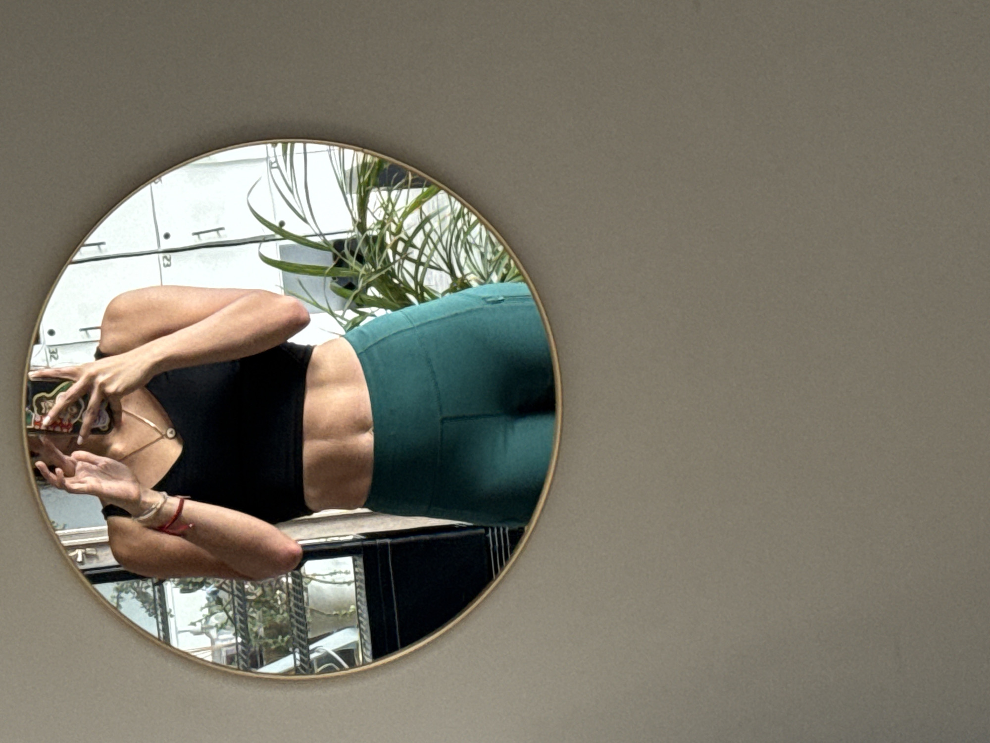 A woman is visible in a round mirror that is hanging on a light grey wall. The woman is positioned on the left side of the mirror, wearing a black top and green leggings. She is holding a phone in her right hand, apparently taking a picture of herself. The reflection shows an urban outdoor scene. The reflection shows bars from a balcony. The reflection contains an outdoor storage locker or cabinet, and foliage. No other details support a location. No conclusion can be reached regarding the location in the city of Puebla, Mexico.