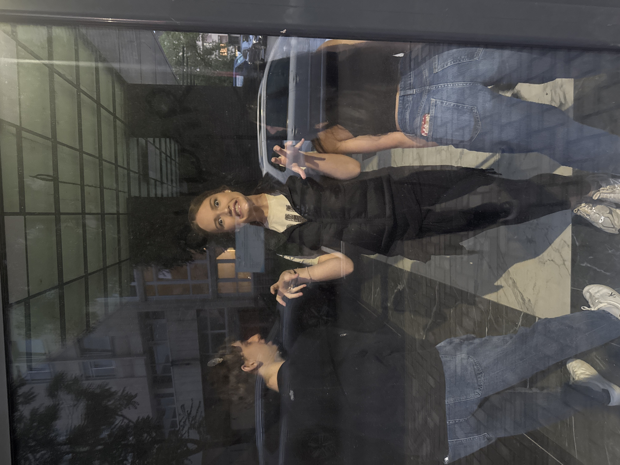 Three individuals are captured in a reflection on a large glass surface, possibly a storefront or building entrance. The scene appears to be outdoors, with a building and parked cars visible in the reflected background.
The person in the center, a young woman, is making a playful, exaggerated facial expression and has her hands raised with fingers splayed, as if mimicking a clawing gesture. She is wearing a dark vest over a light-colored t-shirt and dark pants.
To her left, a young man is seen from the side, wearing a dark sweatshirt and blue jeans. His left arm is extended, and his hand is open.
To the woman's right, another individual, likely a woman given the visible hip and leg area, is facing away from the camera. She is wearing a dark tank top and blue jeans. A small red object is visible in her back pocket.
The ground in the reflection shows a marble-like pattern with geometric shadows cast by overhead structures. The lighting suggests it is either dusk or an overcast day. The overall mood of the image seems informal and playful.