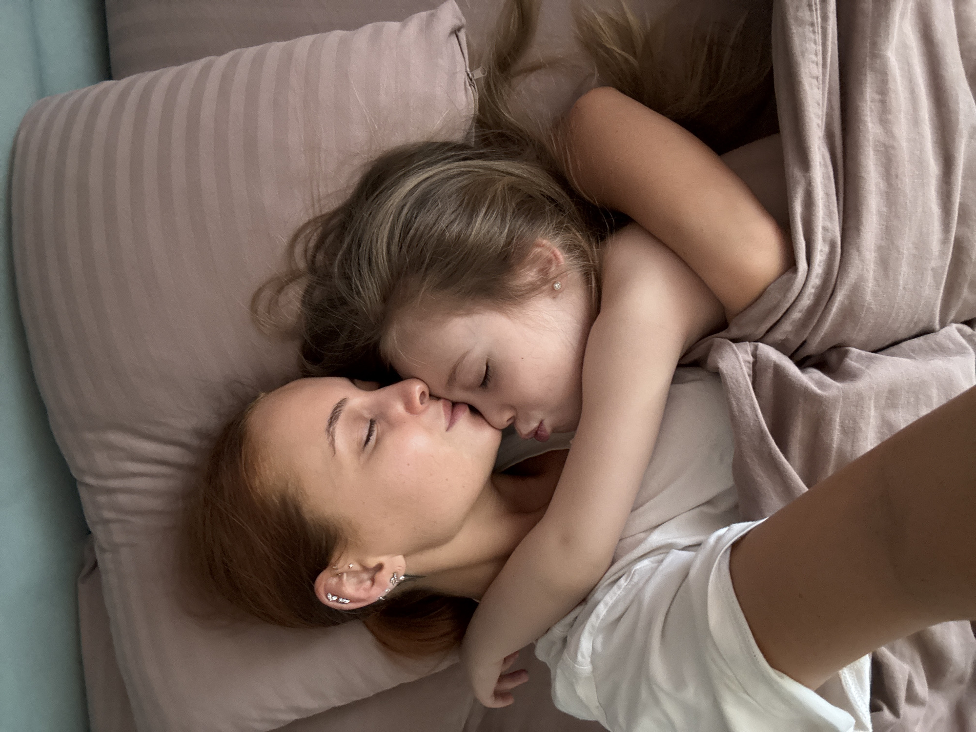 A high-angle shot depicts a woman and a child in bed, likely indoors. The woman, with light red hair and multiple ear piercings, lies on a light pink pillow. A young child, with blonde hair and a single stud earring, rests atop her, their faces close, appearing to kiss. A light brown bed sheet is partially covering them. The woman is wearing a white shirt. The setting is likely a private residence; the exact location within the city of Chaikovsky, Russia cannot be determined from the image.