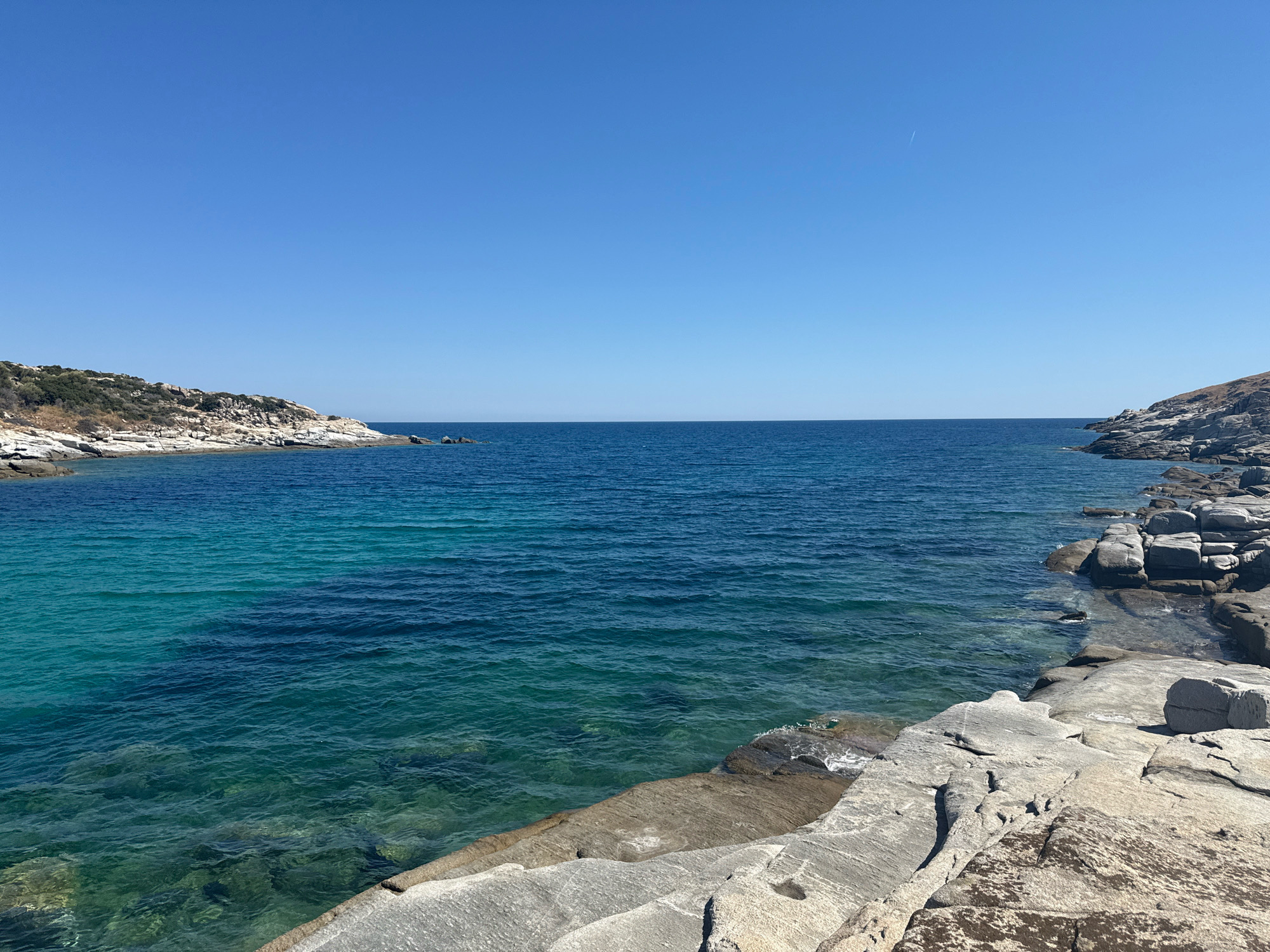 The image depicts a coastal view in Toroni Municipal Unit, Greece, under a clear blue sky. The scene is dominated by the Aegean Sea, with its waters transitioning from a vibrant turquoise near the shore to a deep blue further out. Rocky formations line the coast, with large, light-colored stones forming a natural embankment in the foreground and extending along the right side of the image. To the left and right, hills covered in sparse vegetation rise from the sea. The water is calm, with gentle ripples visible. There are no people or man-made structures immediately apparent in the frame. A faint contrail from an aircraft is visible in the upper right quadrant of the sky.