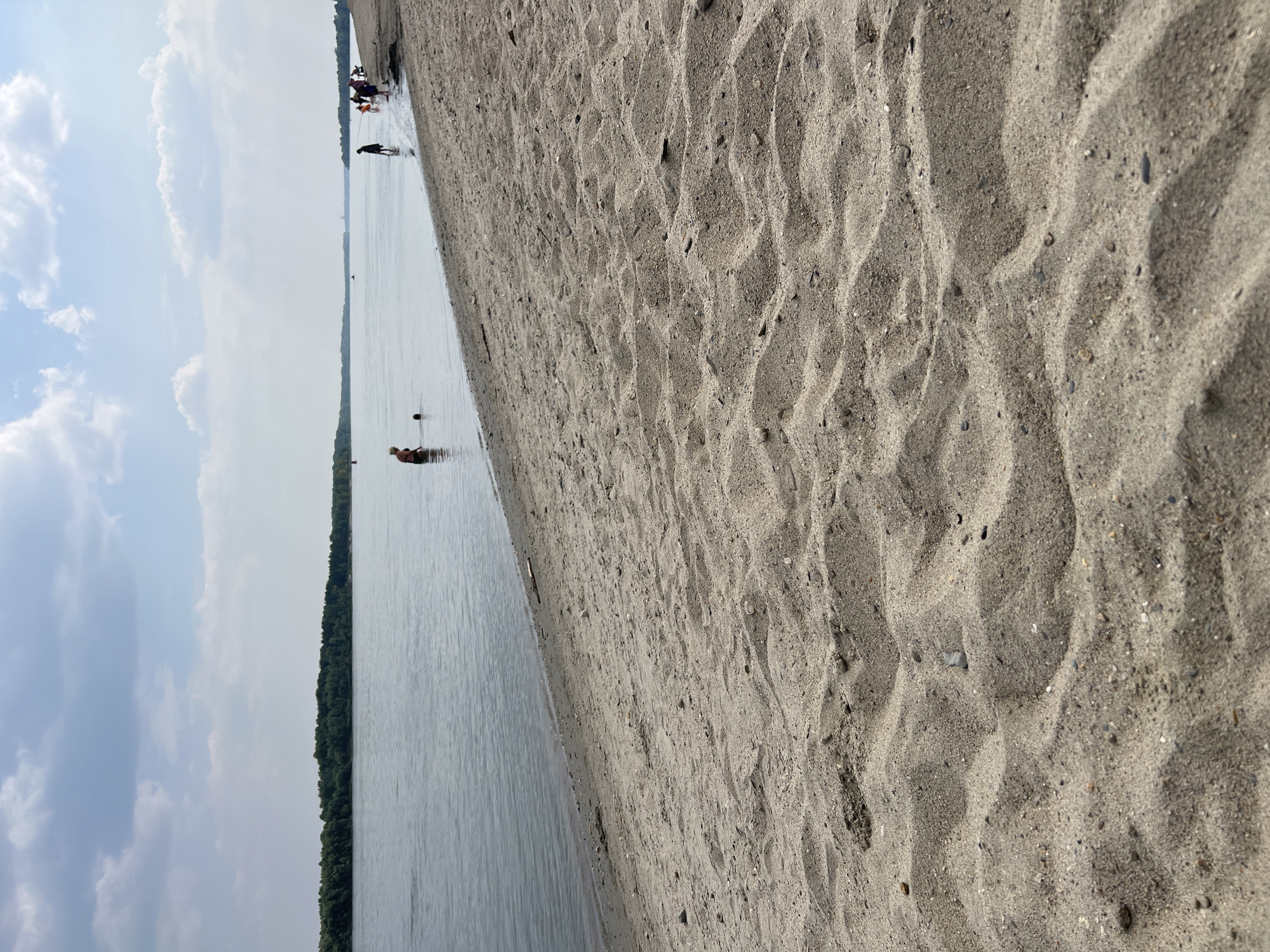 The image depicts a sandy beach along a wide river. The foreground is dominated by textured sand, showing ripples and small pebbles. In the middle ground, the calm river water meets the shore, and several people are visible. Some are wading in the water, while others stand on the beach. A line of trees borders the opposite bank of the river, forming a distant treeline. The sky above is partly cloudy, with scattered white clouds against a light blue backdrop. This scene is consistent with a riverside recreational area, possibly within or near the city of Mochishche, Russia, given its geographical context.