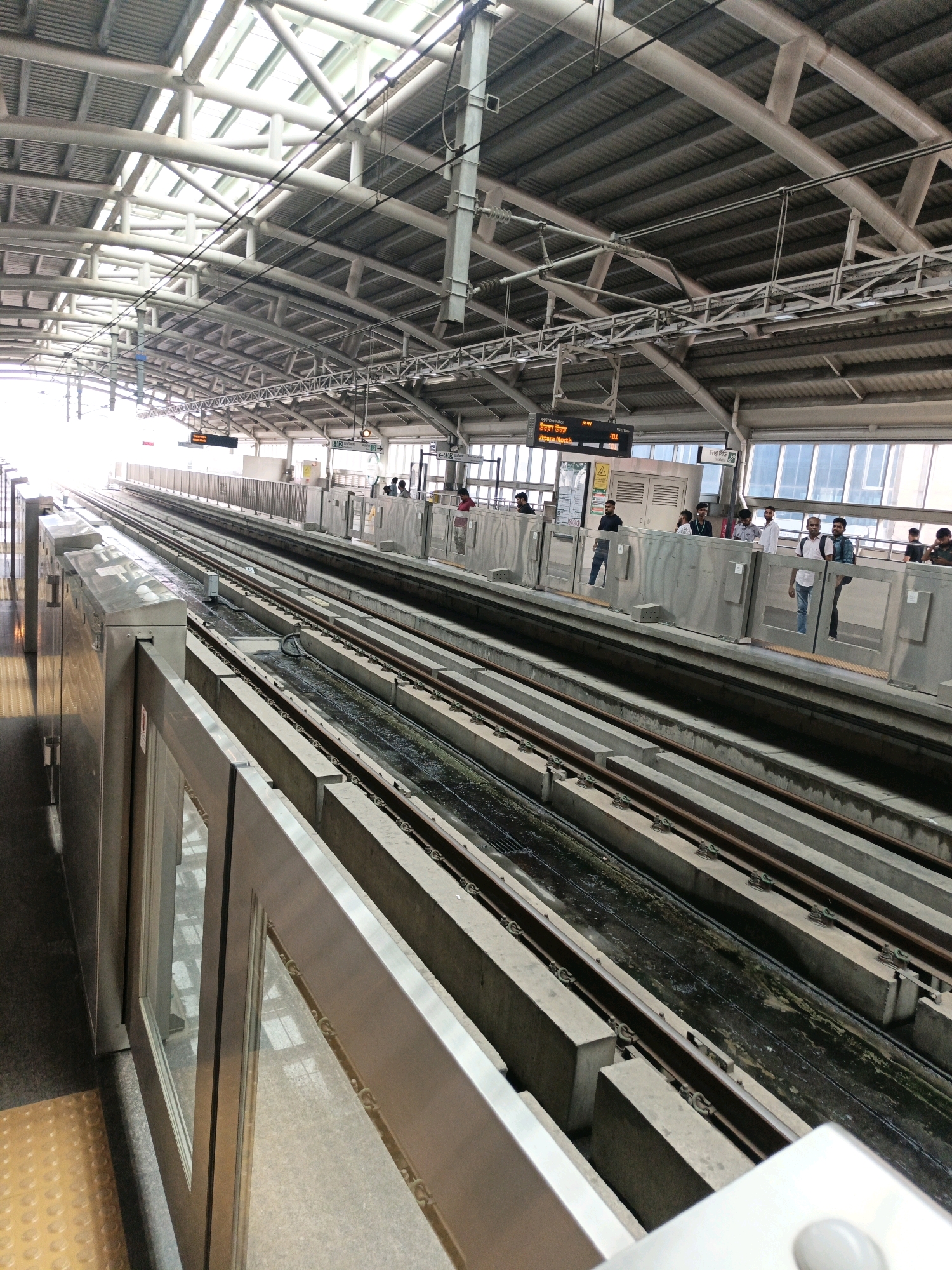 The image shows a view of an elevated metro train station in Dhaka, Bangladesh. The station features multiple tracks, concrete platforms, and a metal roof structure supported by beams. Passengers are visible standing on the platform, awaiting trains. Digital displays show train information. The environment appears to be an active transportation hub with clear visibility of the train tracks and platform infrastructure.