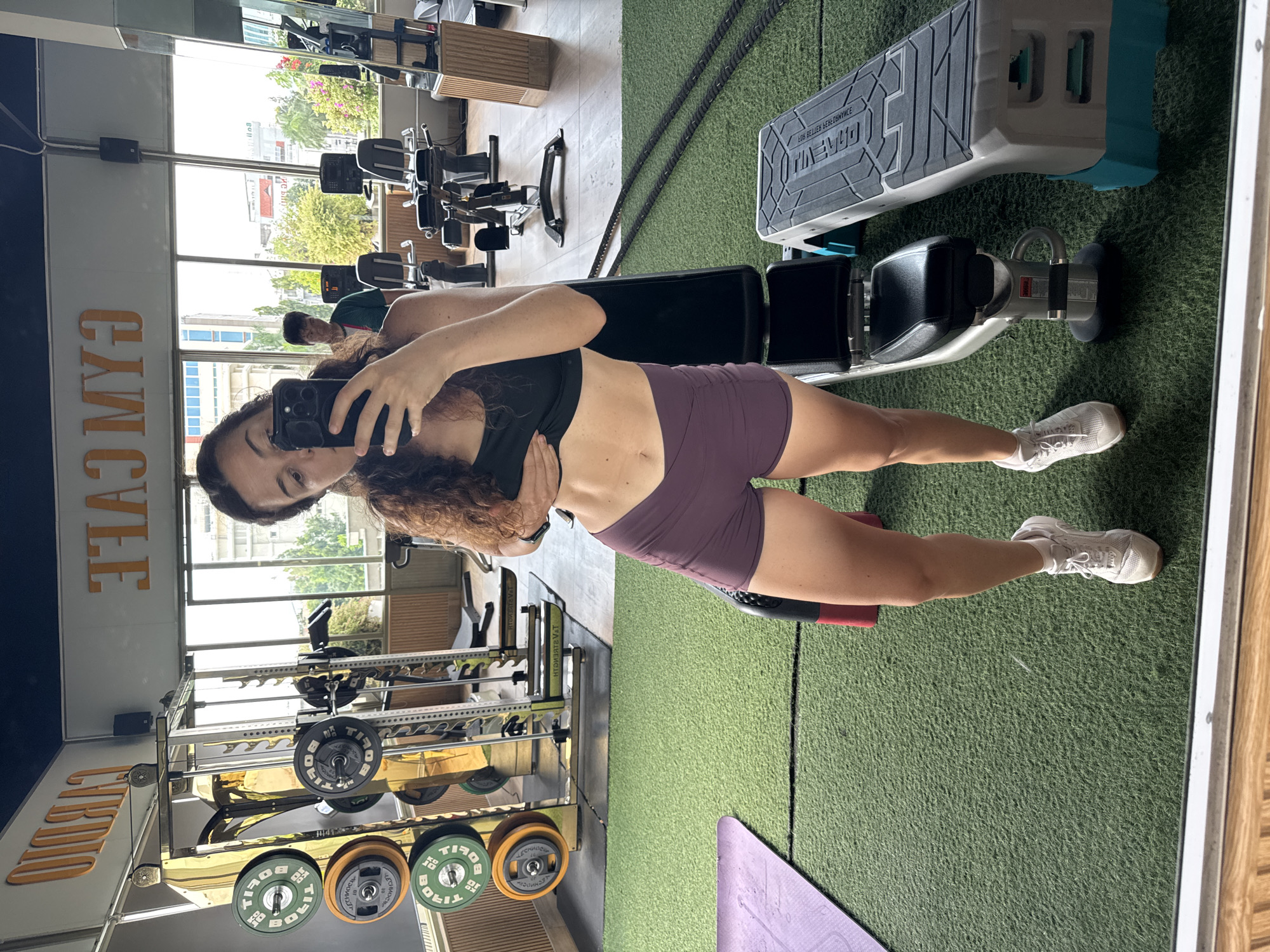 The image shows a woman taking a mirror selfie in a gym located in Phan Thiết, Vietnam. She is wearing a black sports bra and purple athletic shorts, and her abs are visible. The gym features weight racks with weight plates, exercise machines like treadmills, a weight bench, and a plyometric box. The floor is covered with green turf. In the background, through a large window, a cityscape with buildings and trees is visible. A man is also present in the gym, operating an exercise machine. The gym walls have signage that reads "CARDIO" and "GYM CAFE" in orange lettering.
